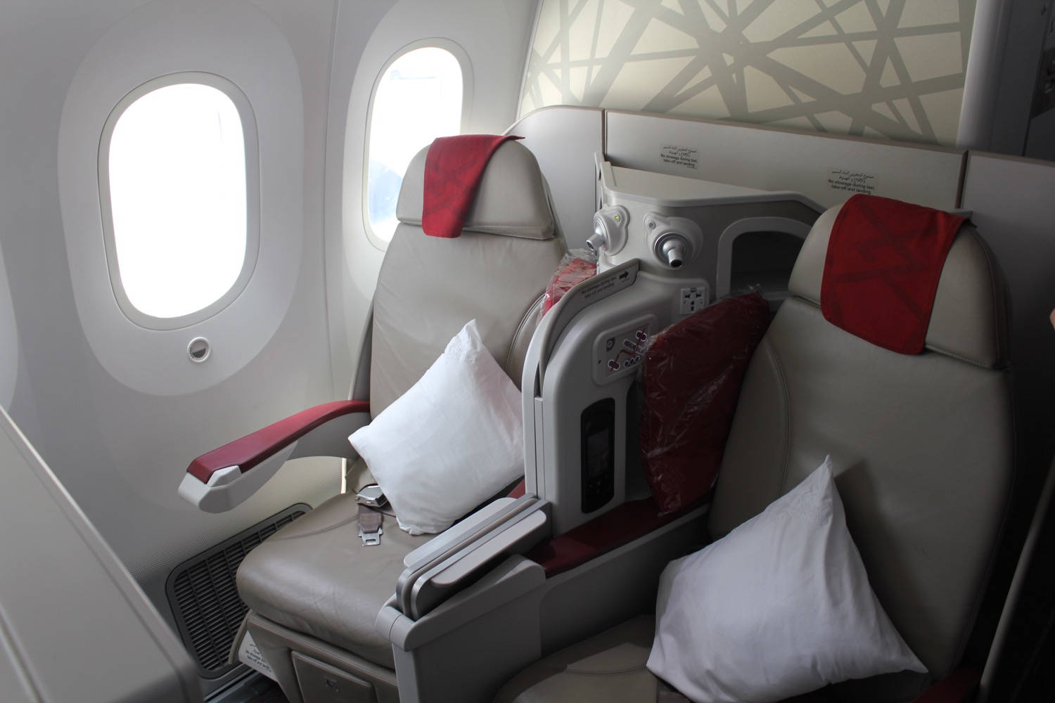 皇家摩洛哥航空飛機後排座椅 壁紙 — 豪華飛機座椅，帶紅色點綴和白色枕頭，位於明亮機艙的橢圓形窗戶旁。