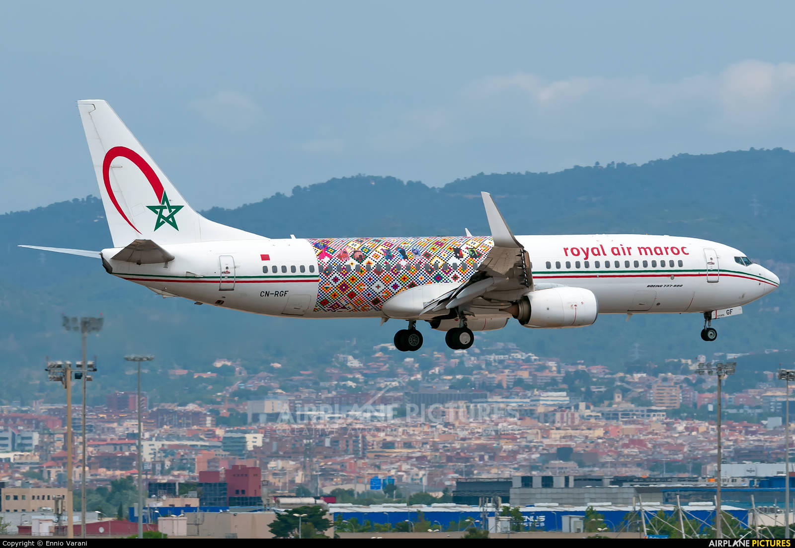 皇家摩洛哥航空飛越城市機隊 壁紙 — 一架摩洛哥皇家航空的波音737，帶有色彩繽紛的幾何圖案塗裝，在煙霧繚繞的山脈背景下，從廣闊的城市景觀上空起飛。