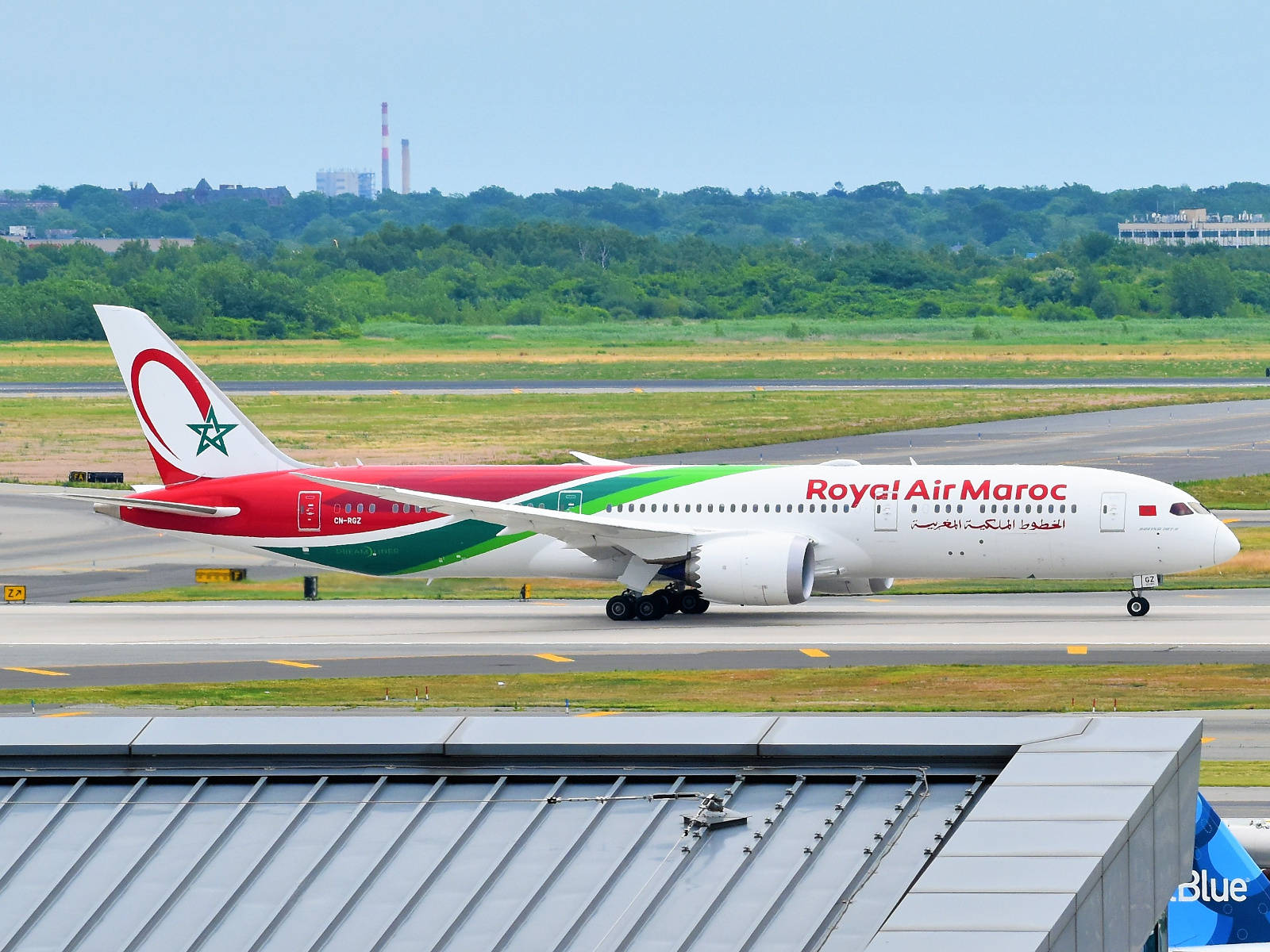 皇家航空馬洛飛機停泊於機場 壁紙 — 一架摩洛哥皇家航空的波音787夢幻客機在陽光明媚的機場跑道上滑行，背景有綠樹和遠處的建築物。