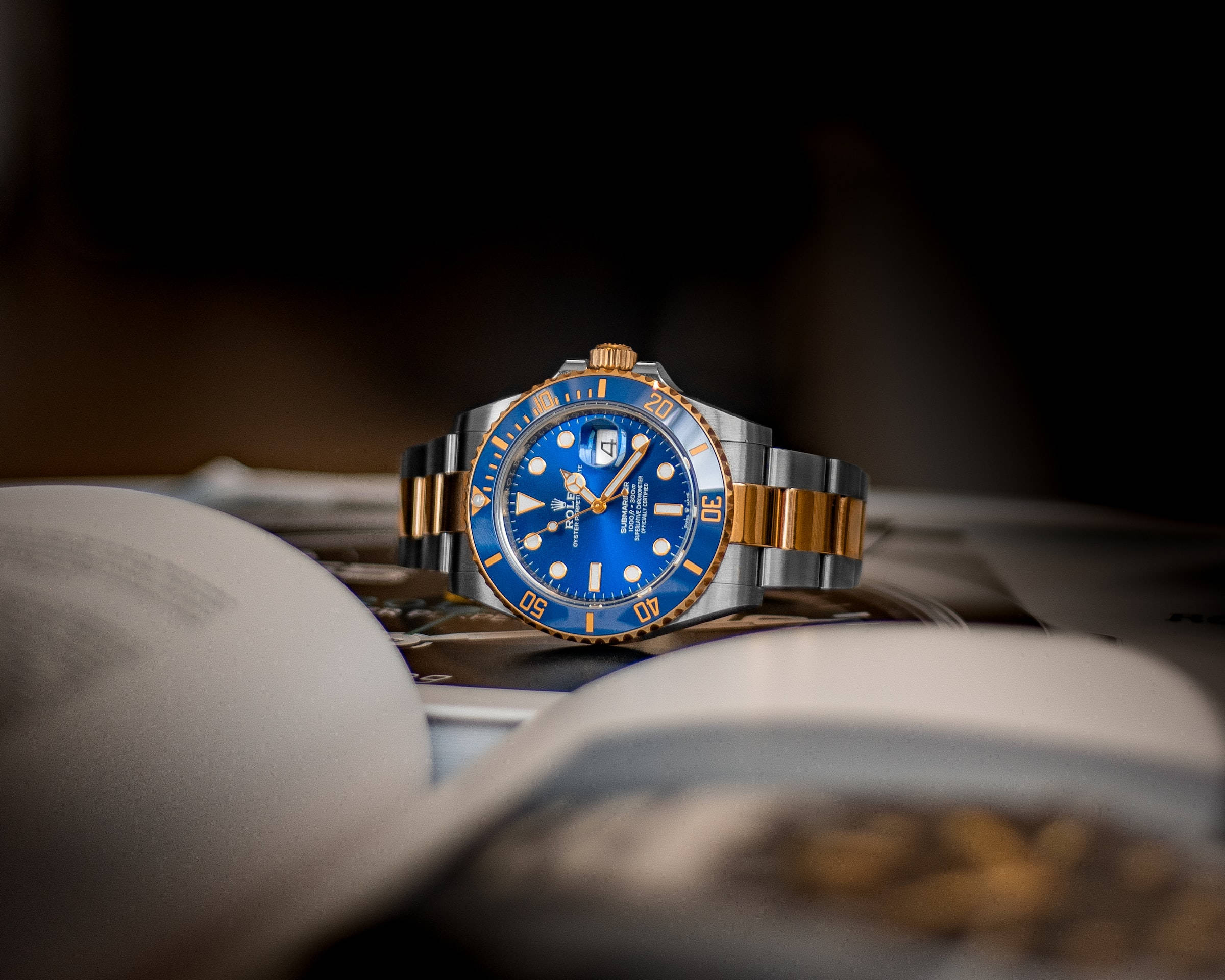 皇家藍勞力士 壁紙 — 一枚豪華的勞力士Submariner，藍色錶盤配金色點綴，優雅地擱在一本打開的書上，在柔和的工作室燈光下。