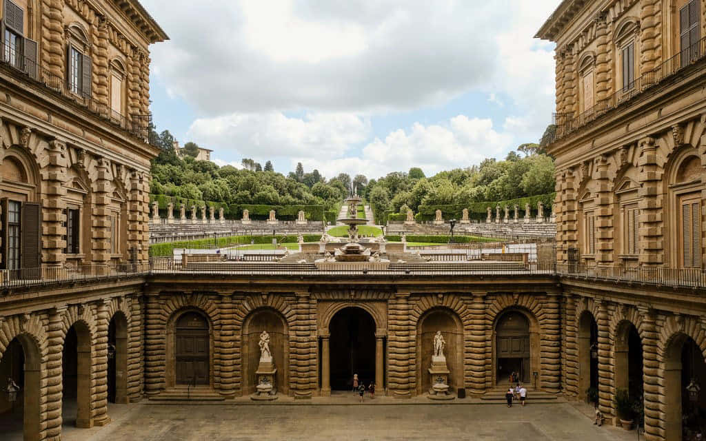 皮蒂宮建築 壁紙 — 宏偉的石頭庭院，周圍環繞著帶有拱形步道和雕像的華麗建築，通向一片在部分多雲天空下的茂密綠色花園。