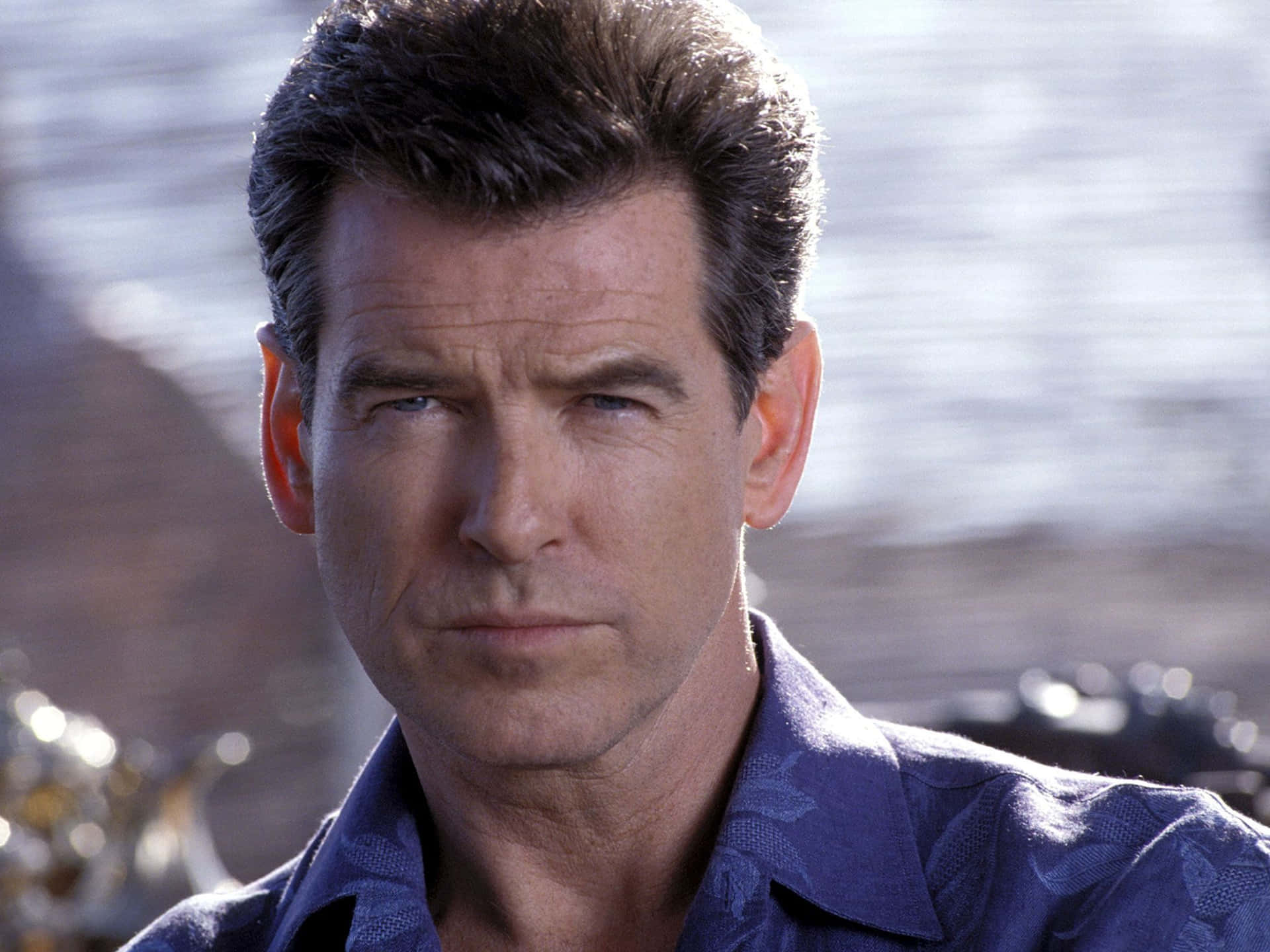 pierce brosnan 桌布 壁紙 — 皮雅斯·布士南的特寫，擁有強烈的藍色眼睛和深色頭髮，穿著藍色圖案襯衫，背景是模糊的水面。