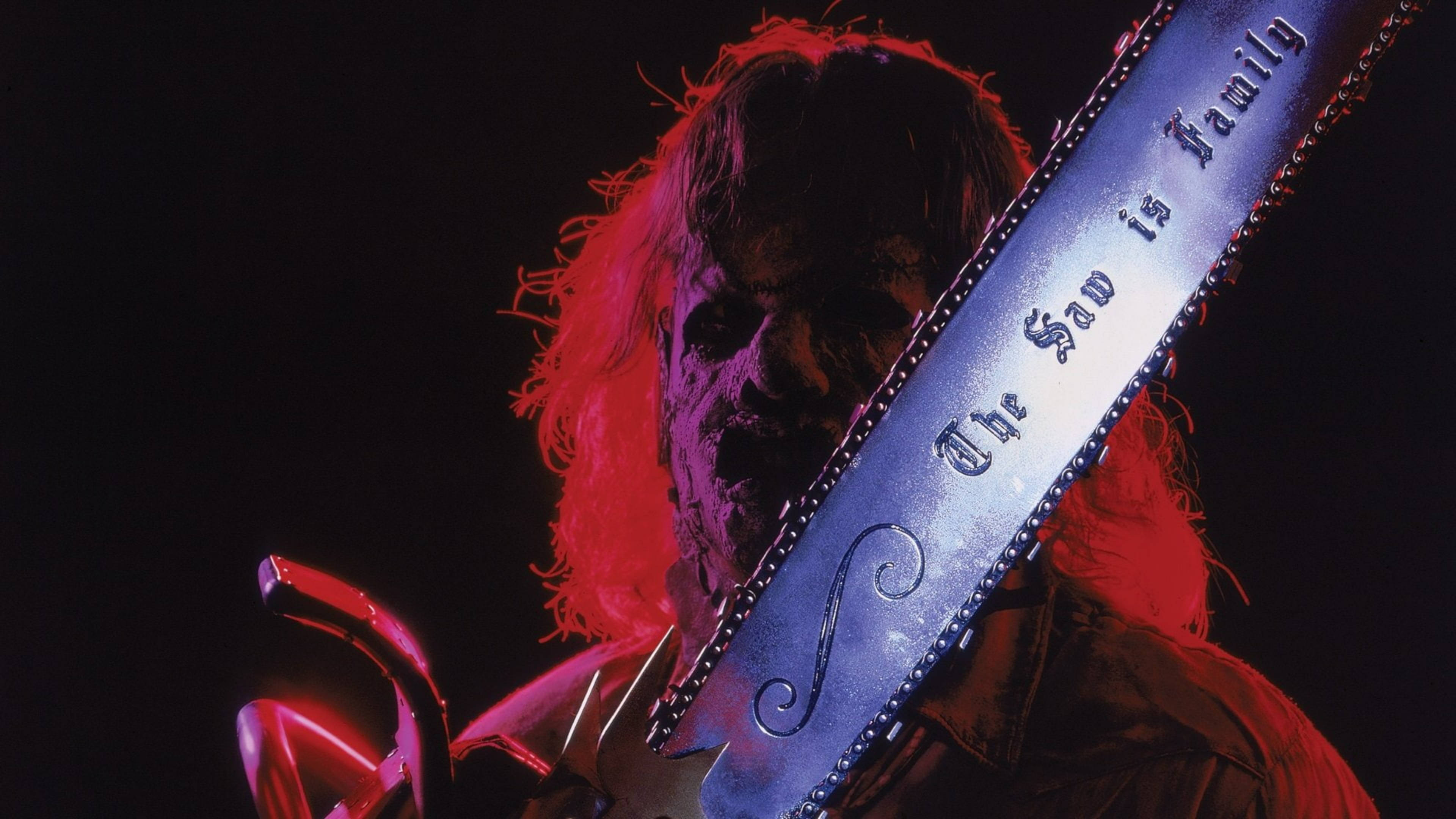皮革臉——《德州電鋸殺人狂III》的驚悚象徵 壁紙 — 一個長紅頭髮的威脅人物，在戲劇性的紅光下拿著一把刻有「The Saw is Family」的電鋸。