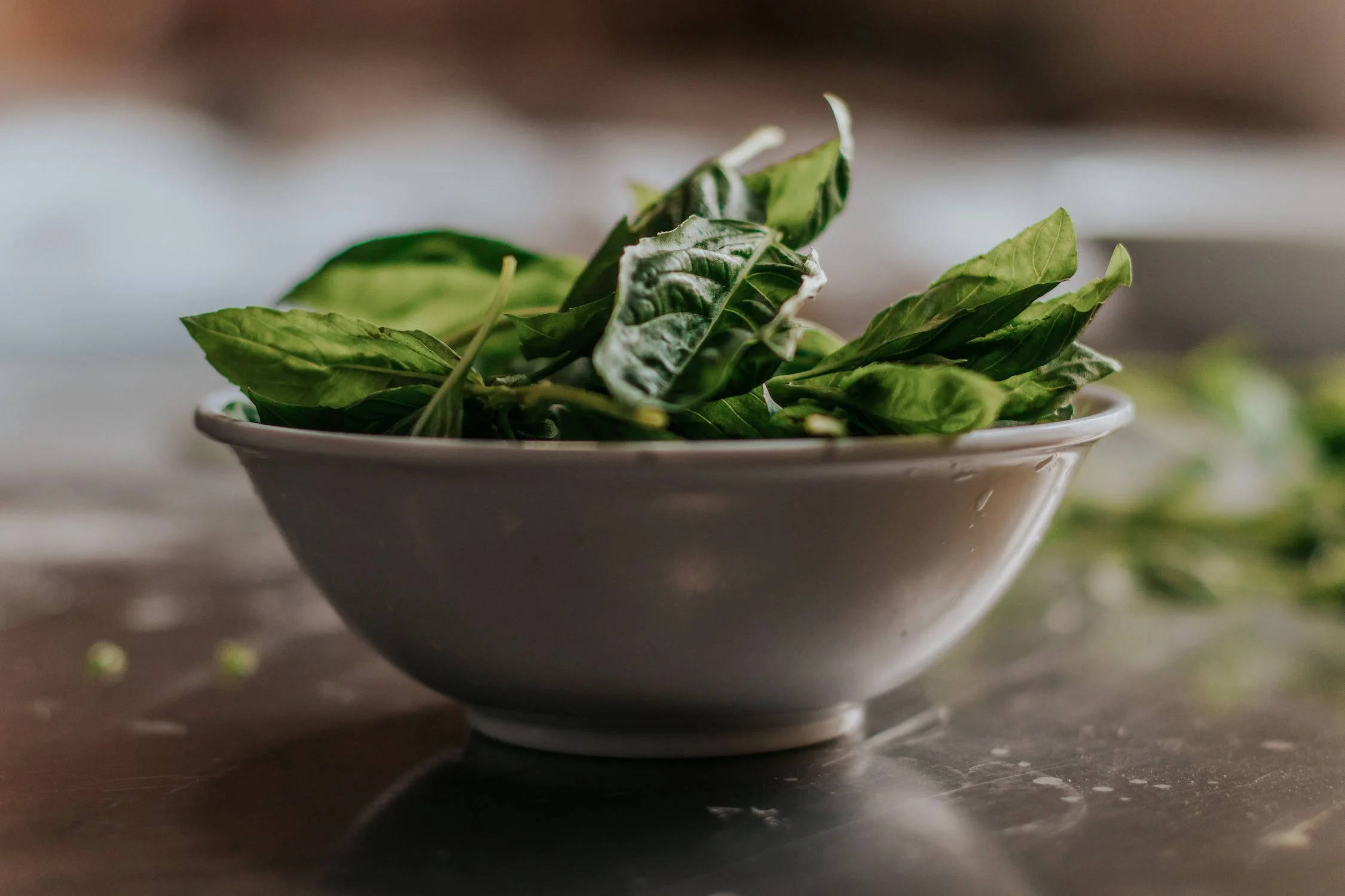 盛於陶瓷碗中的新鮮菠菜 壁紙 — 一個淺白色碗裝滿新鮮綠色羅勒葉，放在深色反光檯面上。