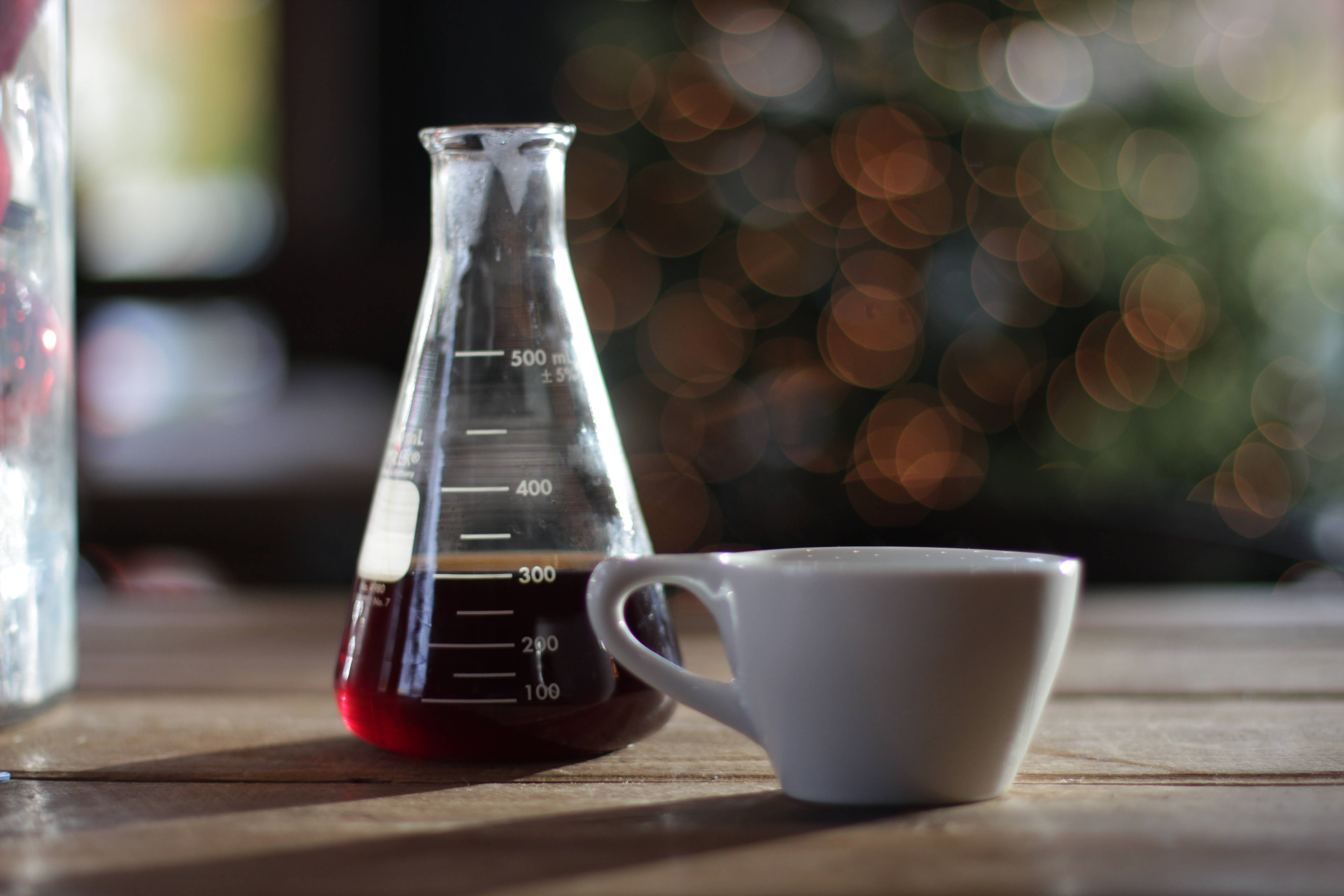 盛有紅色化學品與白杯的燒杯 壁紙 — 一個白色咖啡杯放在木桌上，旁邊是一個裝滿深色咖啡的玻璃燒杯，背景有溫暖的散景燈光。