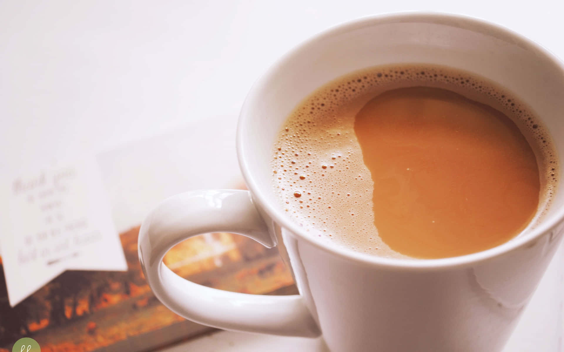盛滿奶油咖啡的茶杯圖 壁紙 — 一個白色馬克杯的特寫，裝滿冒煙的咖啡和泡沫，旁邊有一張感謝卡和一本書，放在明亮的表面上。