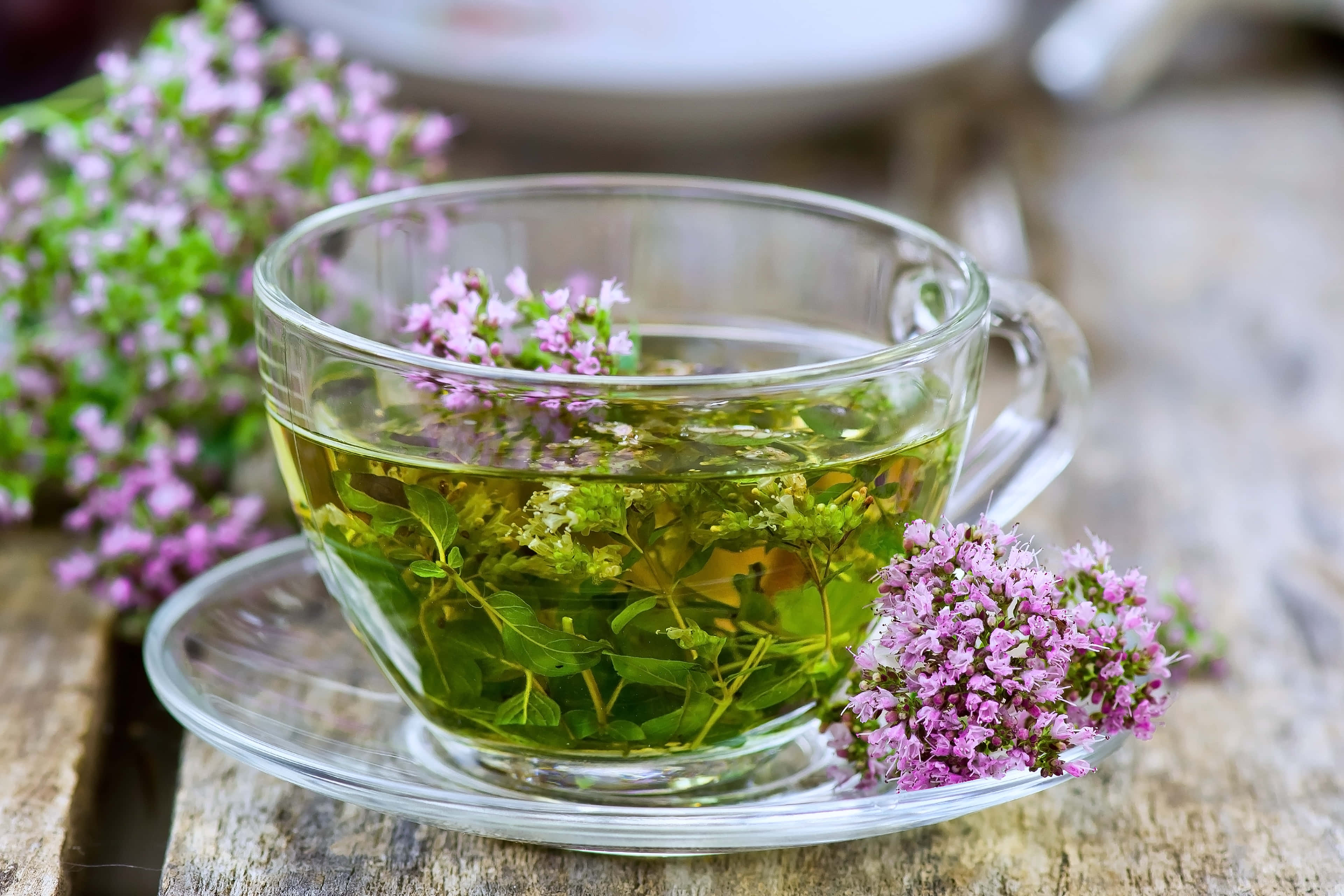 盛滿薄荷葉的茶杯圖 壁紙 — 一個透明玻璃杯盛載著冒煙的草本茶，浮著綠色葉片和紫色花朵，放在質樸的木桌上。