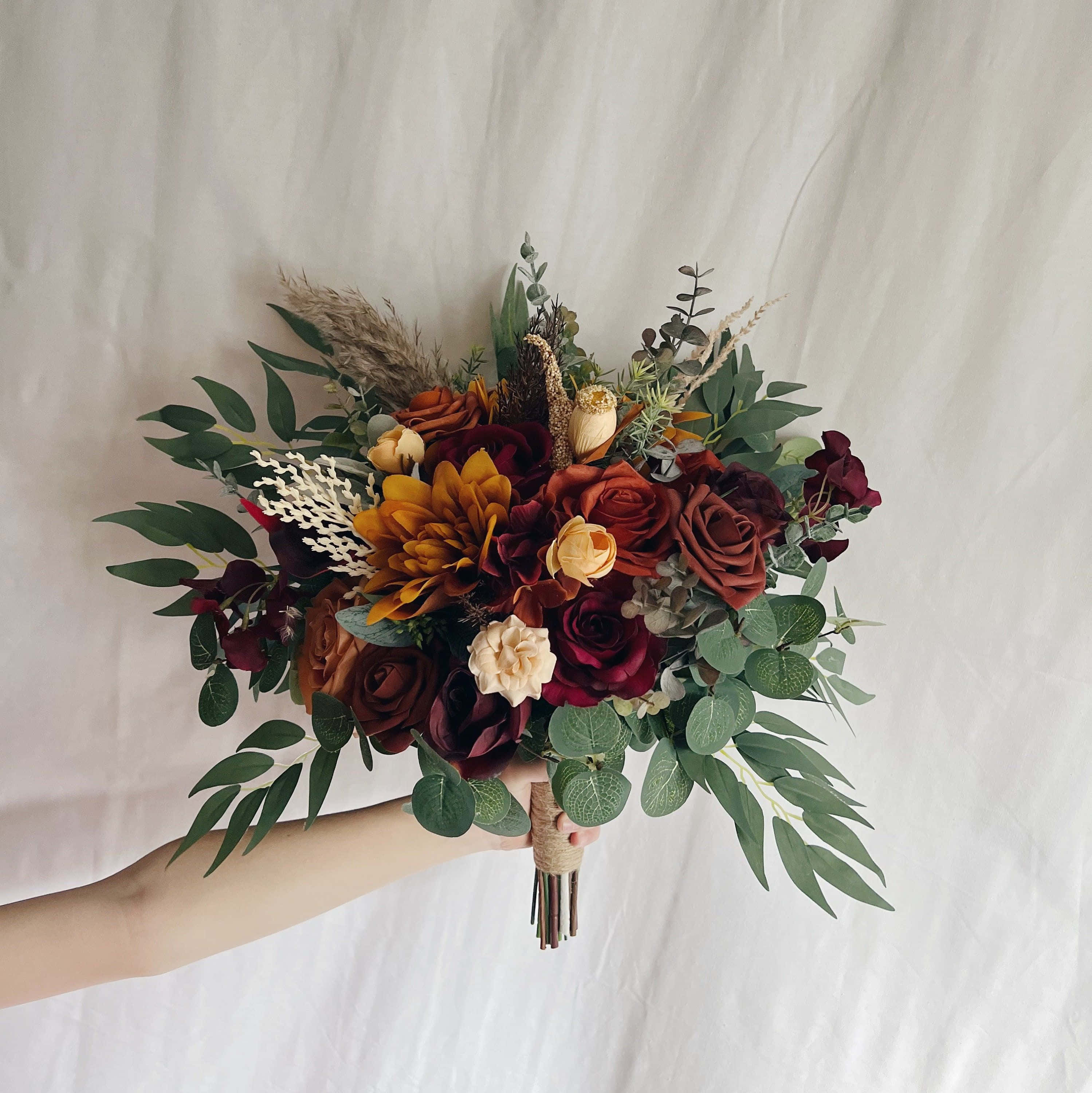 盛開花束圖 壁紙 — 一隻手拿著一束豐盛的秋日花束，包含深紅玫瑰、琥珀色大麗花和綠葉，背景是柔和的白色。