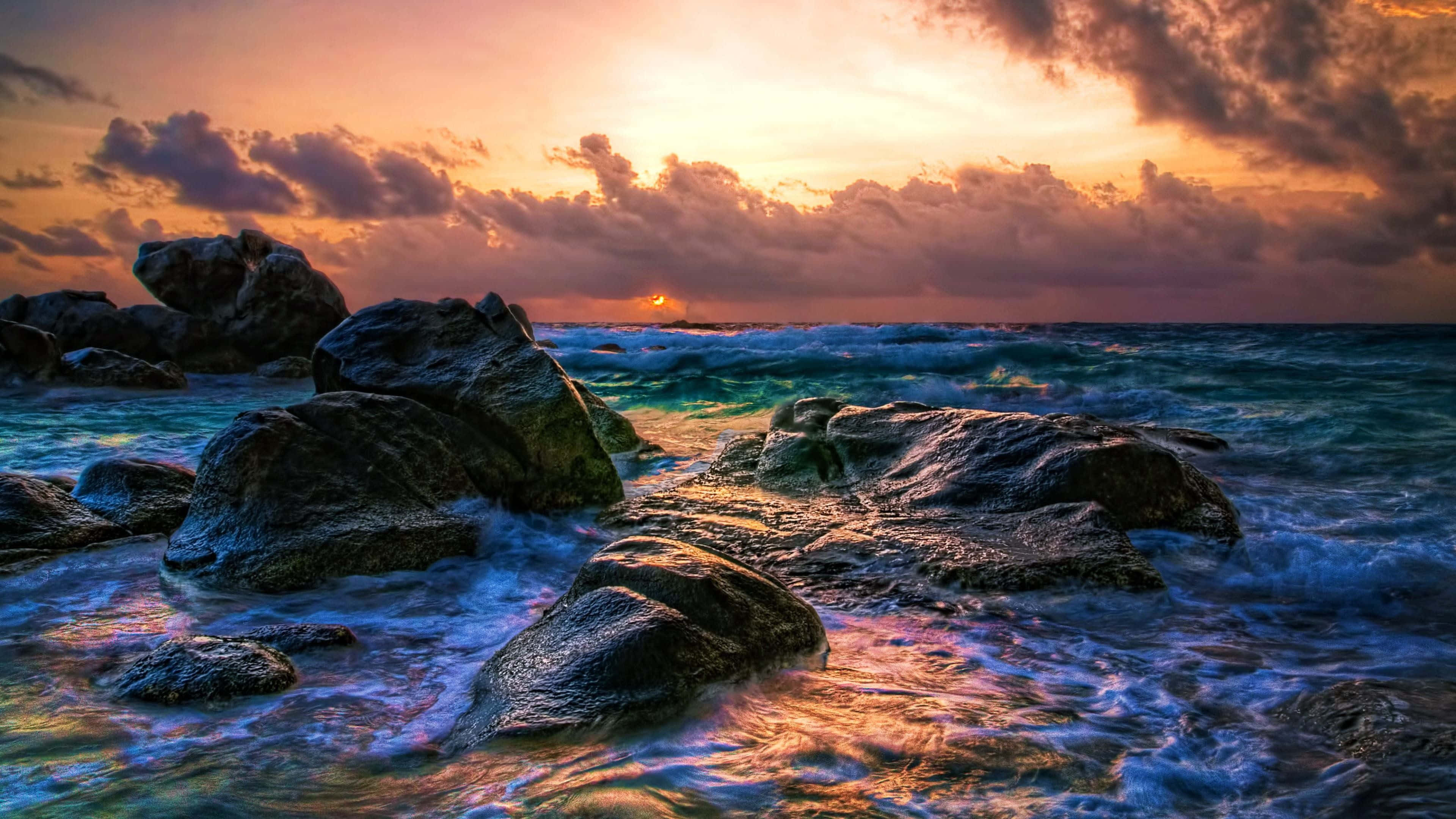 盡享 UHD 美景 壁紙 — 夕陽將天空染成火熱的橙色和紫色，海浪拍打著海岸的黑色岩石。