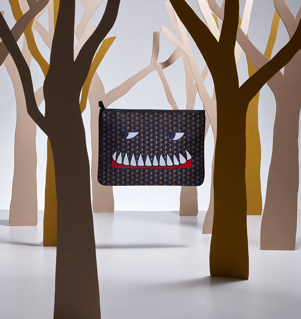 盡享奢華：Moynat Bad Dreams 手拿包 壁紙 — 一個棕色圖案的手拿包，帶有卡通怪物面孔，懸掛在簡約工作室環境中的風格化紙樹之間。