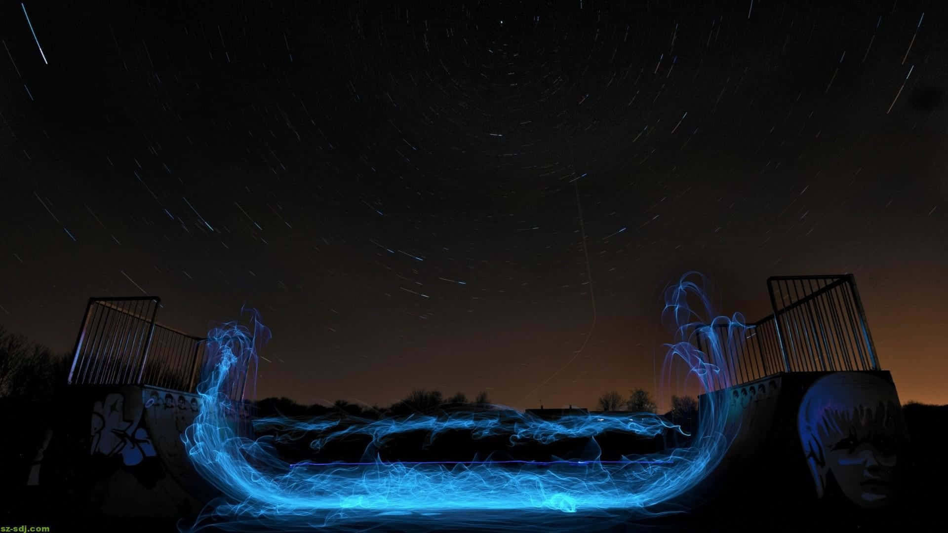 盡享這款鮮豔的1080p AMOLED背景 壁紙 — 一條發光的藍色光跡在星空下跨越黑暗的橋樑，可見星軌。
