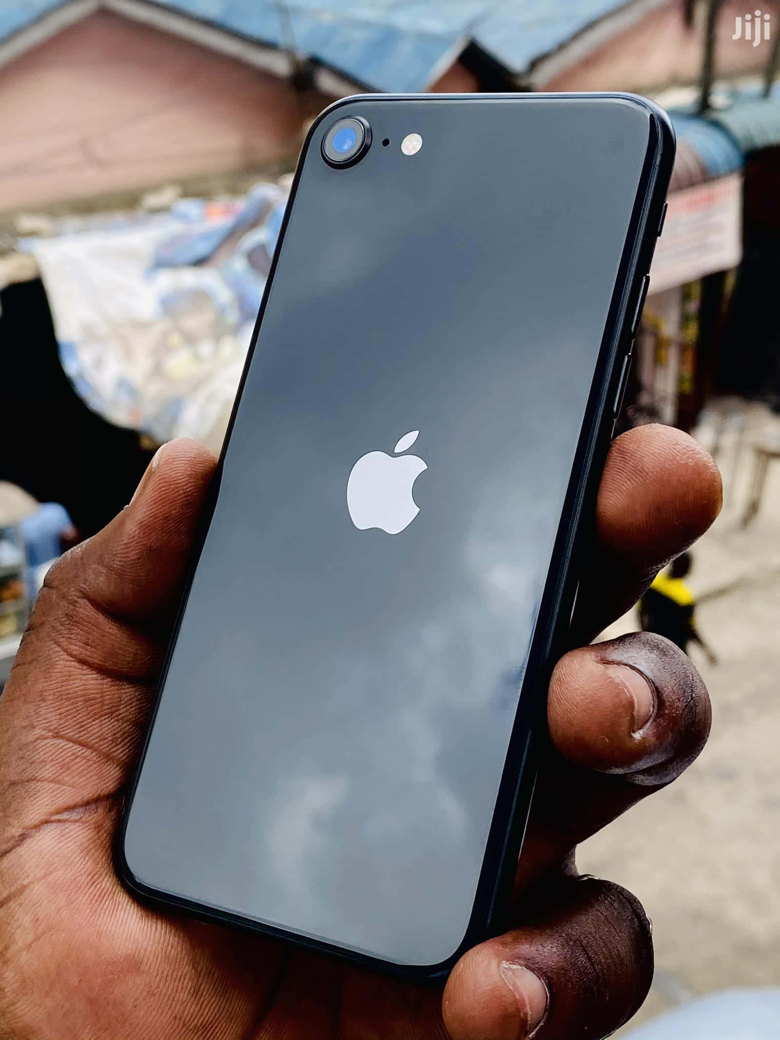 盡情享受你的最新Apple手機 壁紙 — 一隻手拿著一部黑色iPhone，背面反光顯示多雲的天空，背景是模糊的戶外市場。