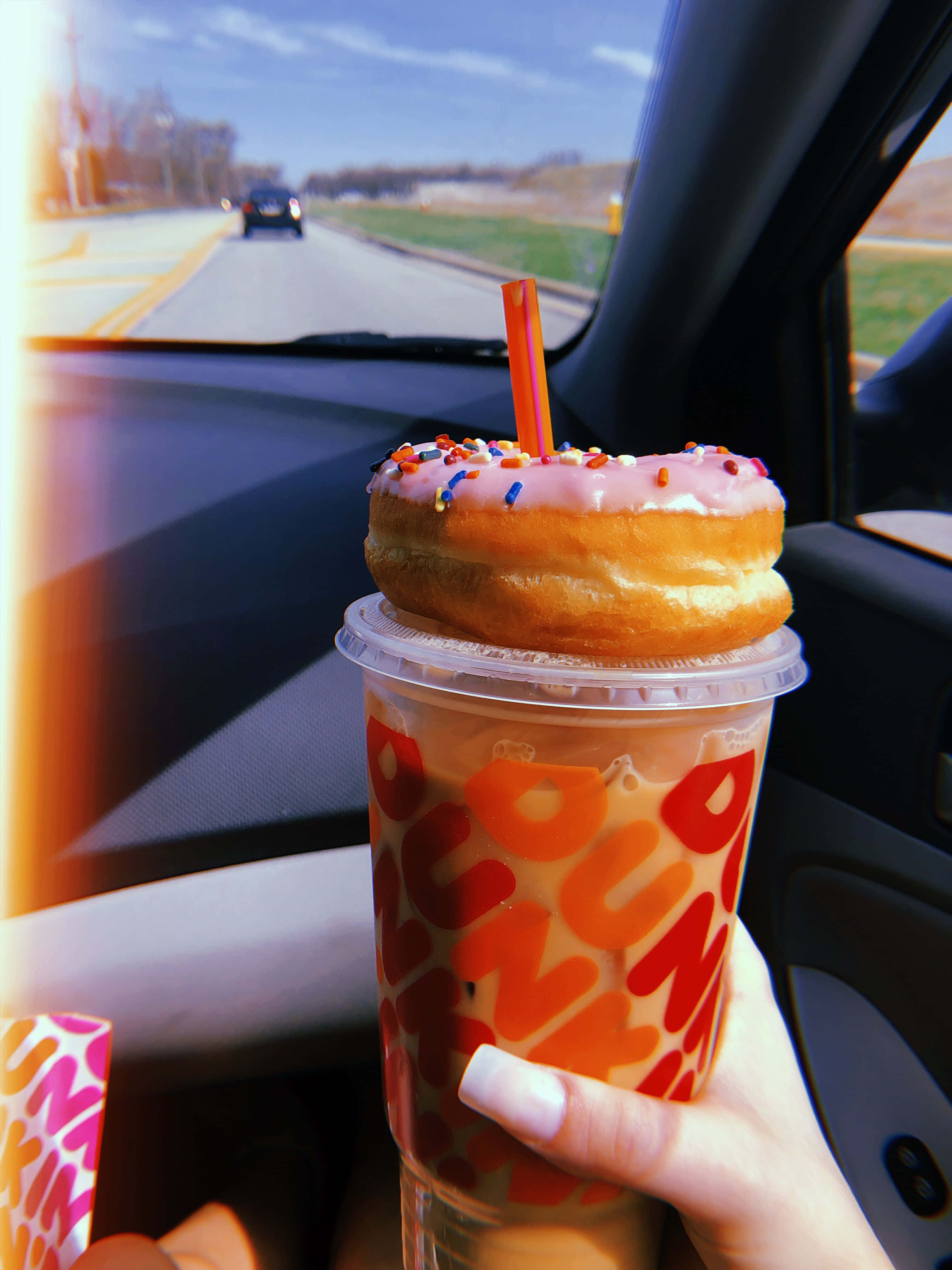 盡情享用Dunkin' Donuts 壁紙 — 在陽光明媚的公路旅行日，一隻手在車內拿著一杯頂部有粉紅色糖霜甜甜圈和糖粒的Dunkin'冰咖啡。