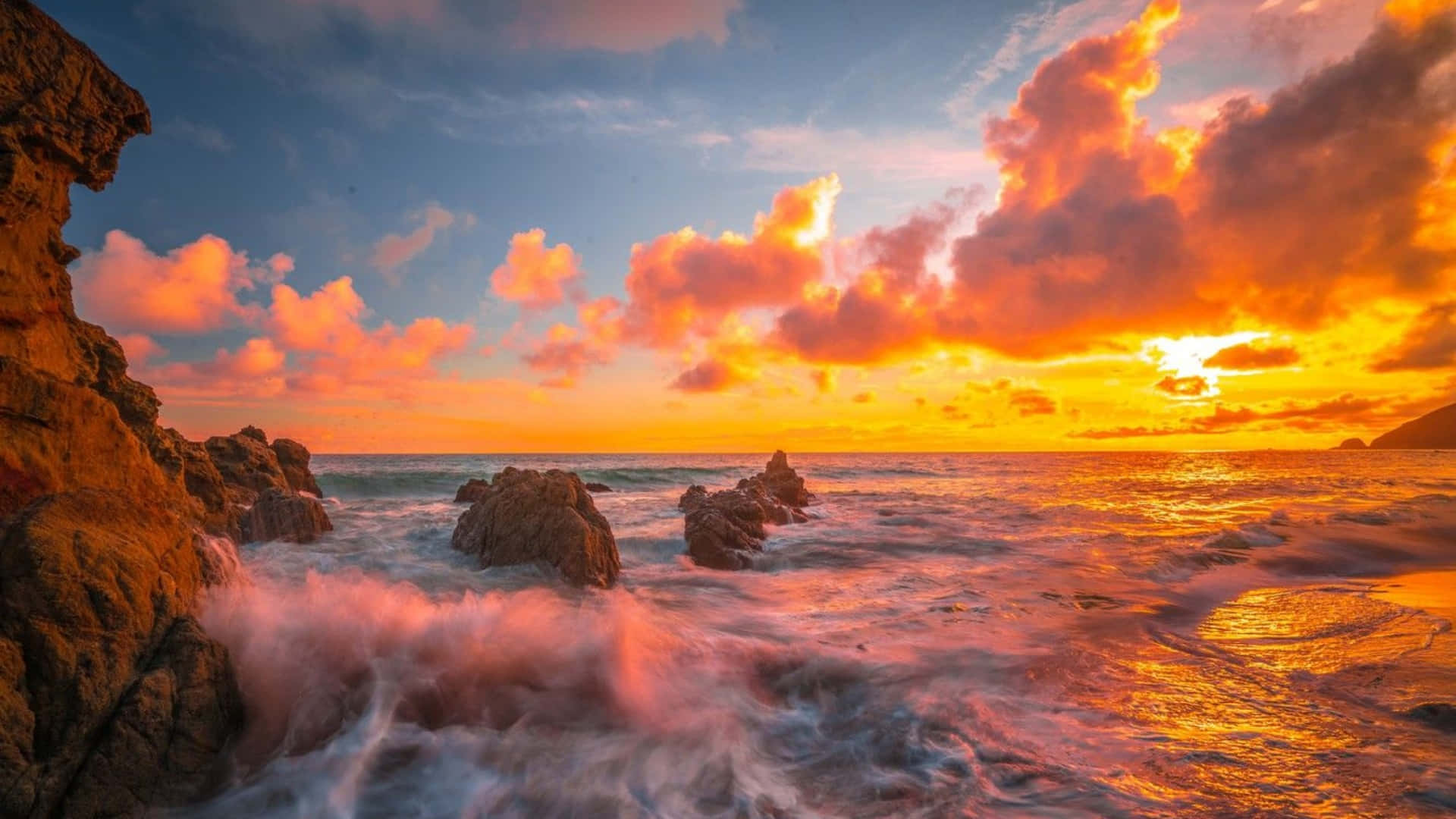 盡情欣賞加州馬利布的絕美風光 壁紙 — 夕陽將天空染成火熱的橙色和粉紅色，海浪拍打著崎嶇的海岸岩石。
