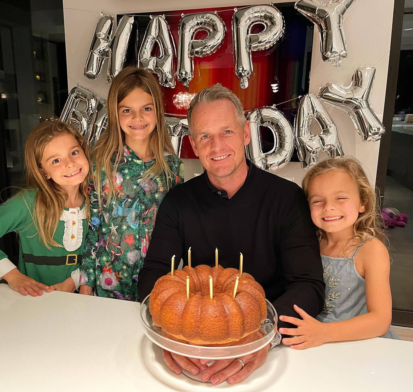 盧克·唐納德慶祝生日 壁紙 — 一名男子和三個微笑的女孩在銀色「HAPPY BIRTHDAY」氣球下圍著一個插有蠟燭的環形蛋糕擺姿勢。