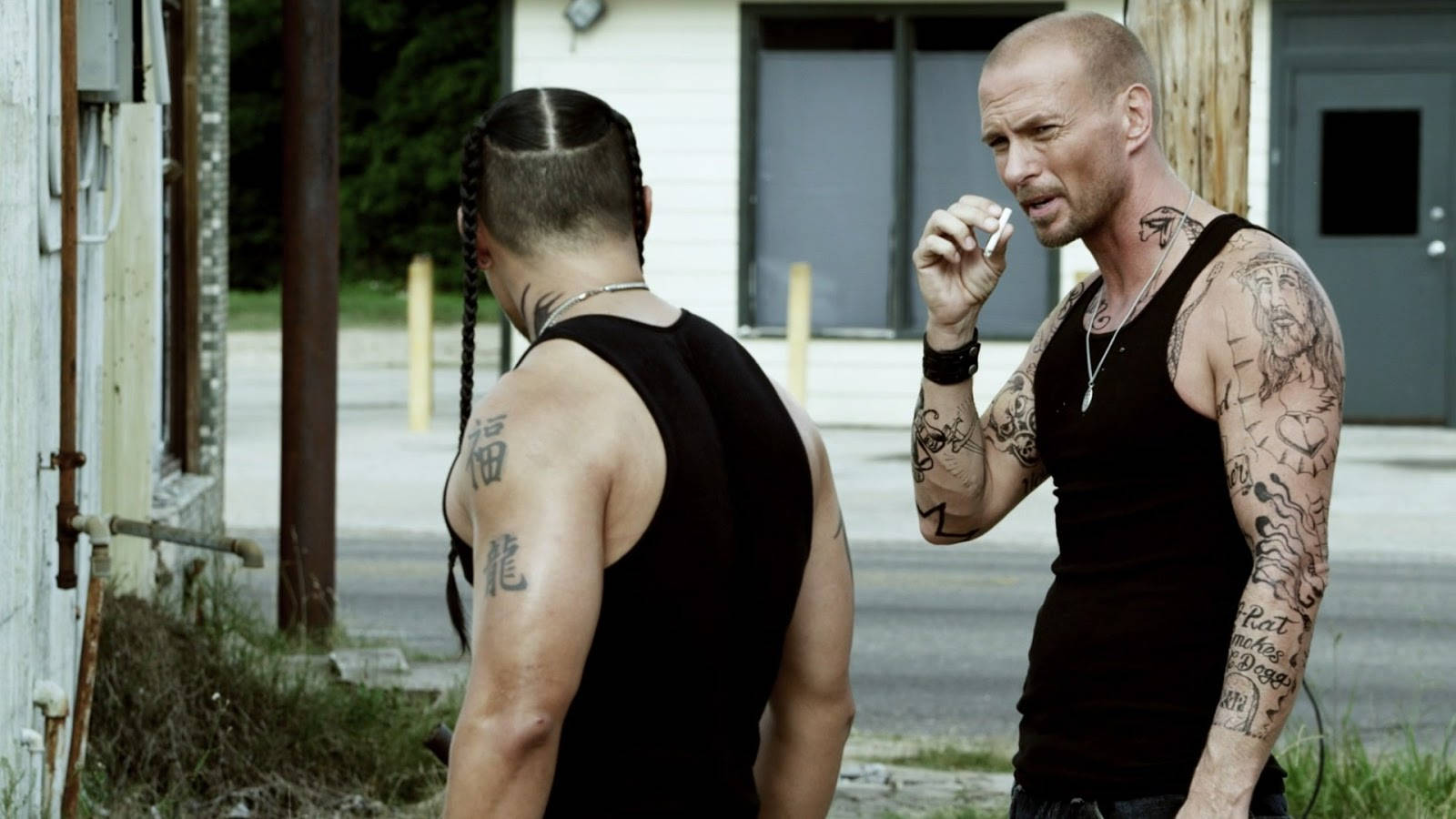 盧克·高斯紋身肌膚 壁紙 — 兩名紋身男子身穿黑色背心，在一條雜草叢生、建築物破舊的骯髒城市街道上對峙。