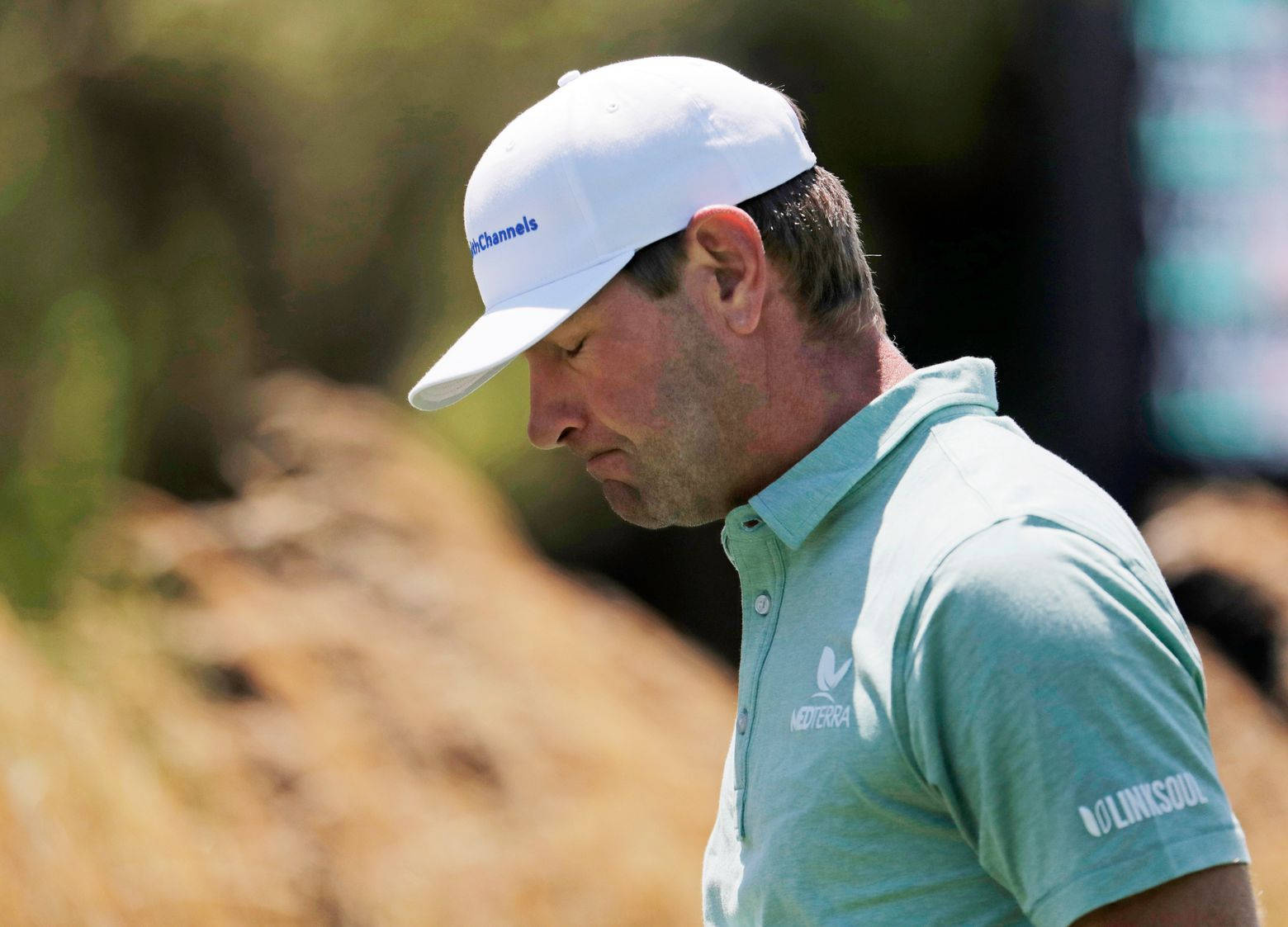 盧卡斯·格洛弗顯露沮喪神情 壁紙 — 一位專注的高爾夫球手，戴著白色帽子，身穿淺藍色Polo衫，在陽光明媚的球場上專注地低頭看著。