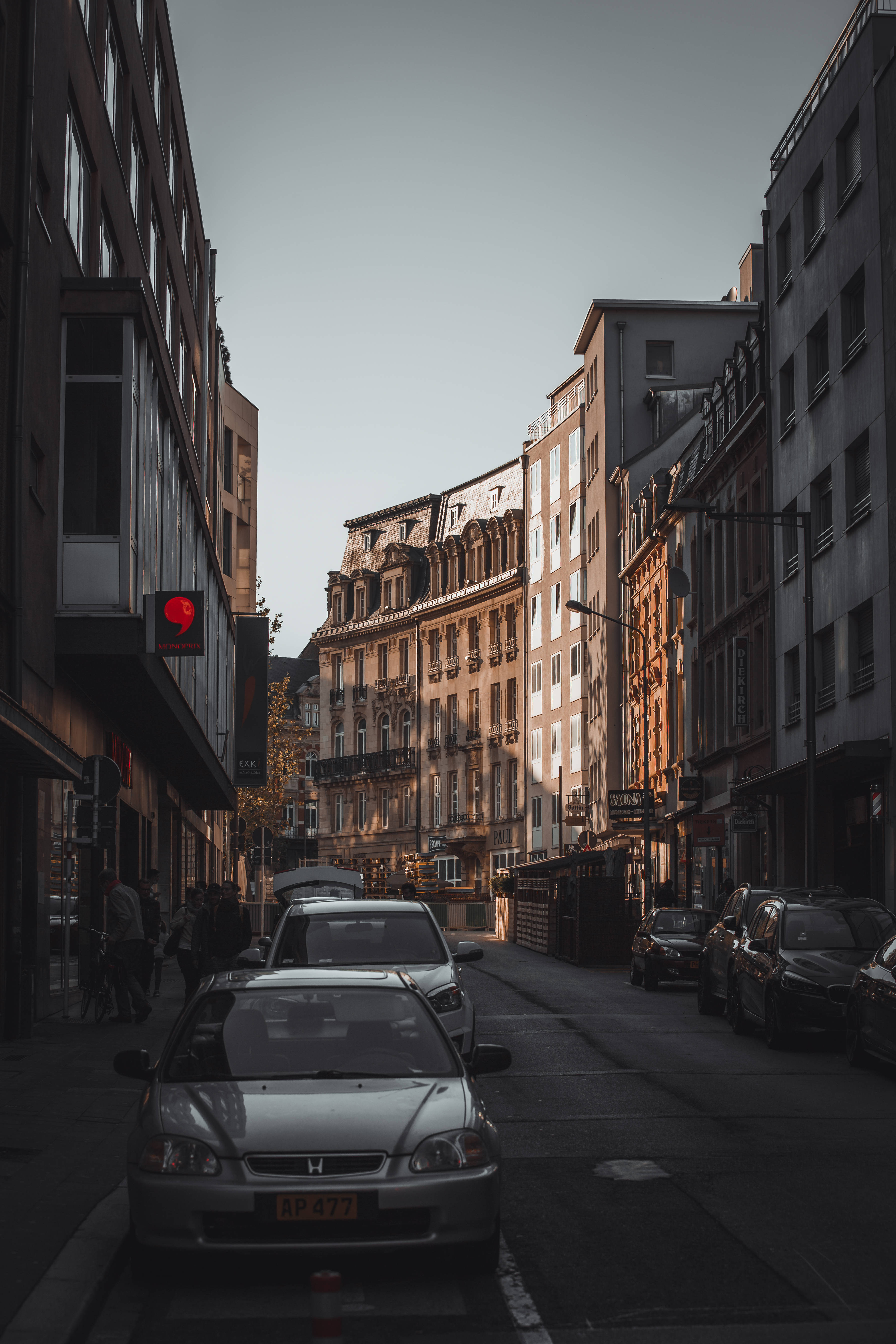 盧森堡市街景 壁紙 — 一條狹窄的城市街道，兩旁停滿汽車和高樓大廈，沐浴在柔和的金色時刻光線中。