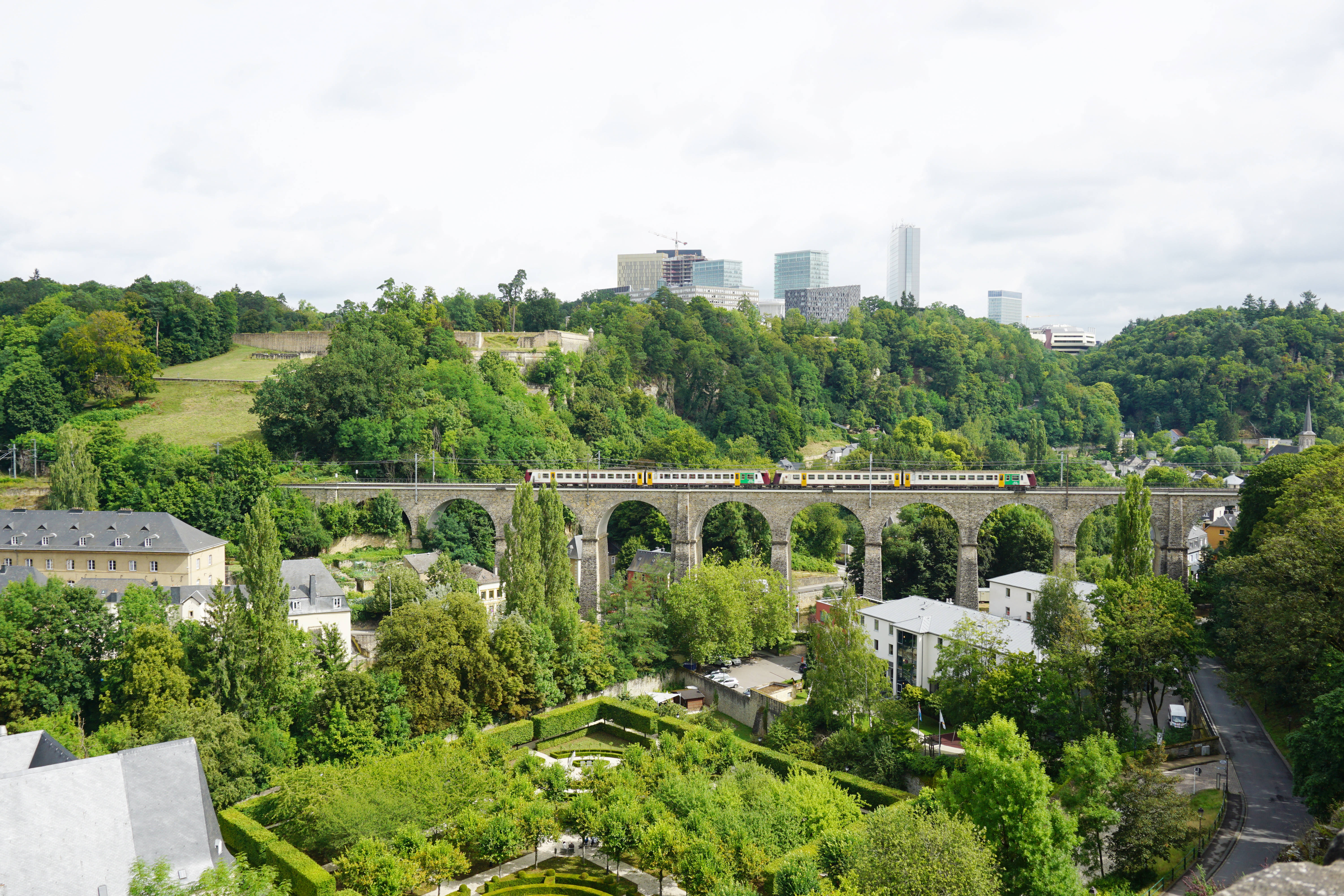 盧森堡拉帕塞勒橋 壁紙 — 一列火車穿過石拱橋，下方是鬱鬱蔥蔥的花園，現代城市建築在樹木覆蓋的山丘後方升起，天空多雲。
