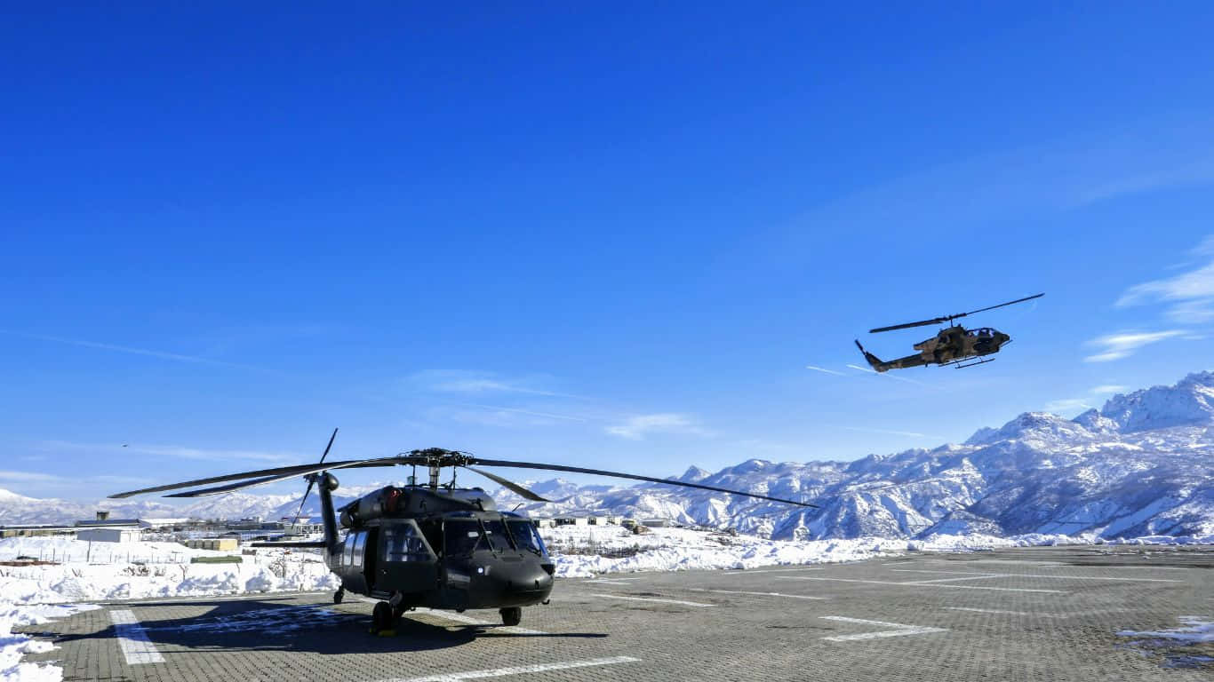 直升機隊飛越雪山的空中視角 壁紙 — 兩架黑色軍用直升機，一架停泊在積雪的停機坪上，另一架在晴朗的藍天中飛行，背景是白雪覆蓋的山脈。