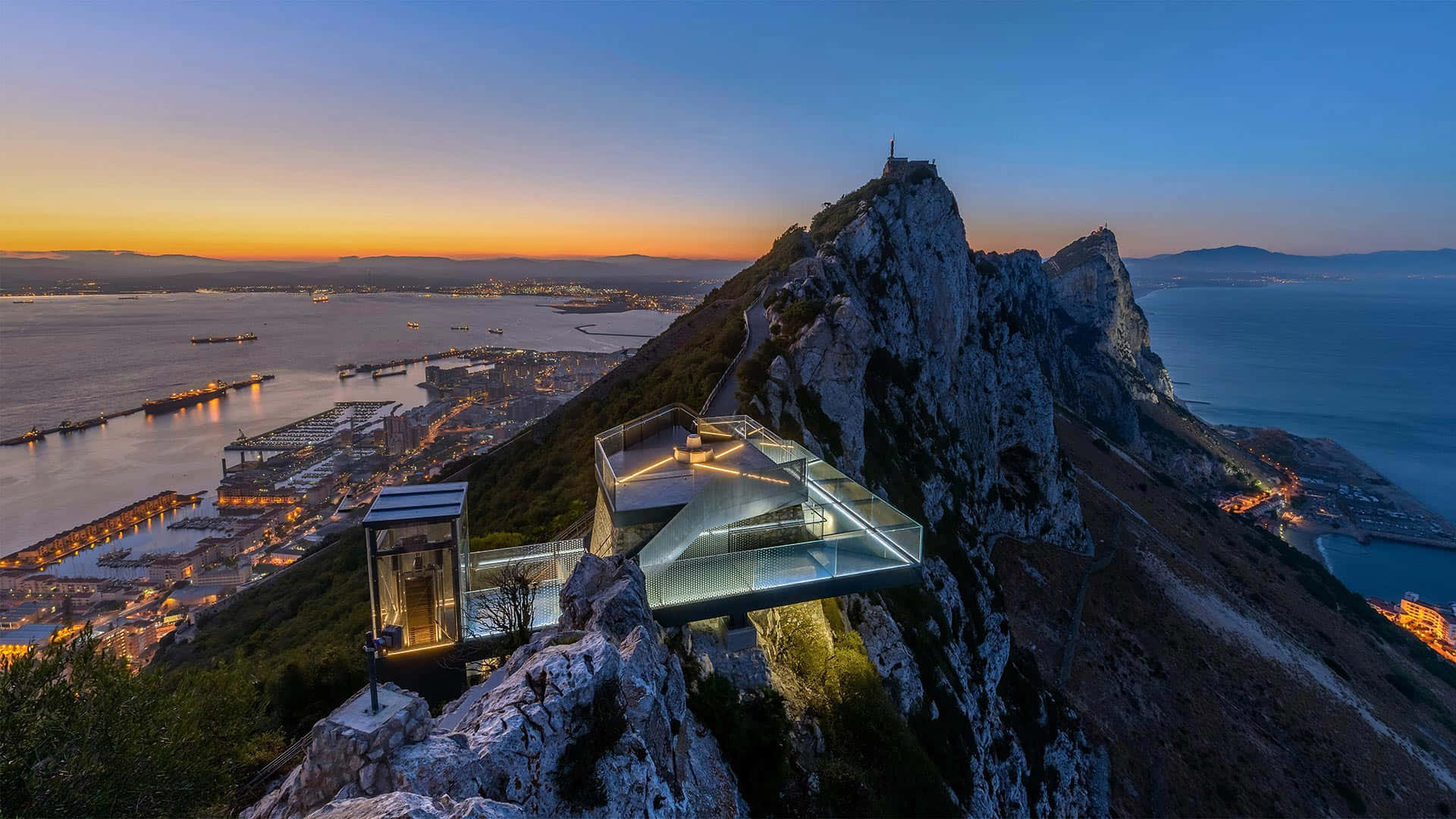 直布羅陀岩夜空美景 壁紙 — 一座玻璃幕牆的現代建築戲劇性地棲息在岩石懸崖上，在日落時分俯瞰沿海城市和港口。