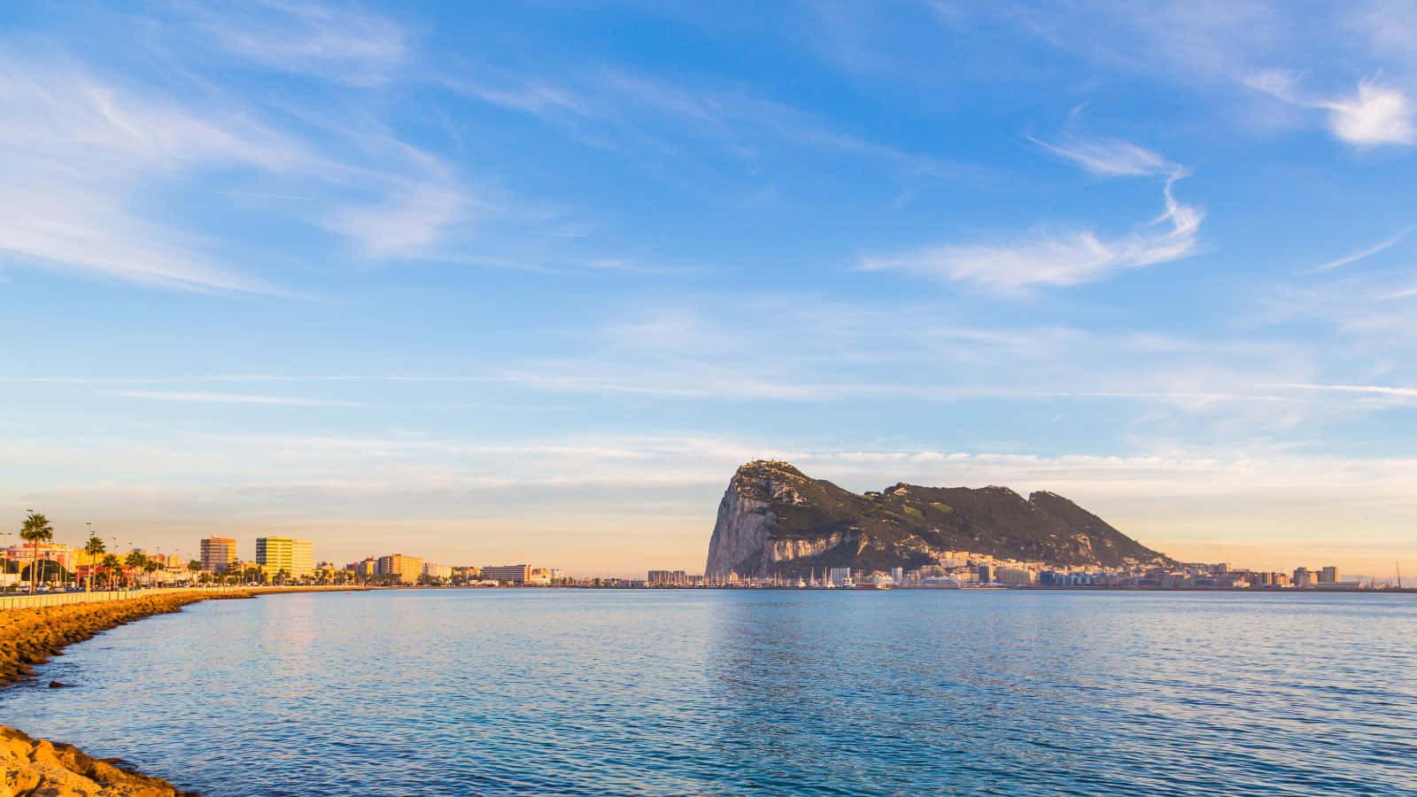 直布羅陀岩海景 壁紙 — 直布羅陀巨岩在黃金時刻的寧靜海岸景觀，平靜的藍色海水和晴朗的天空。