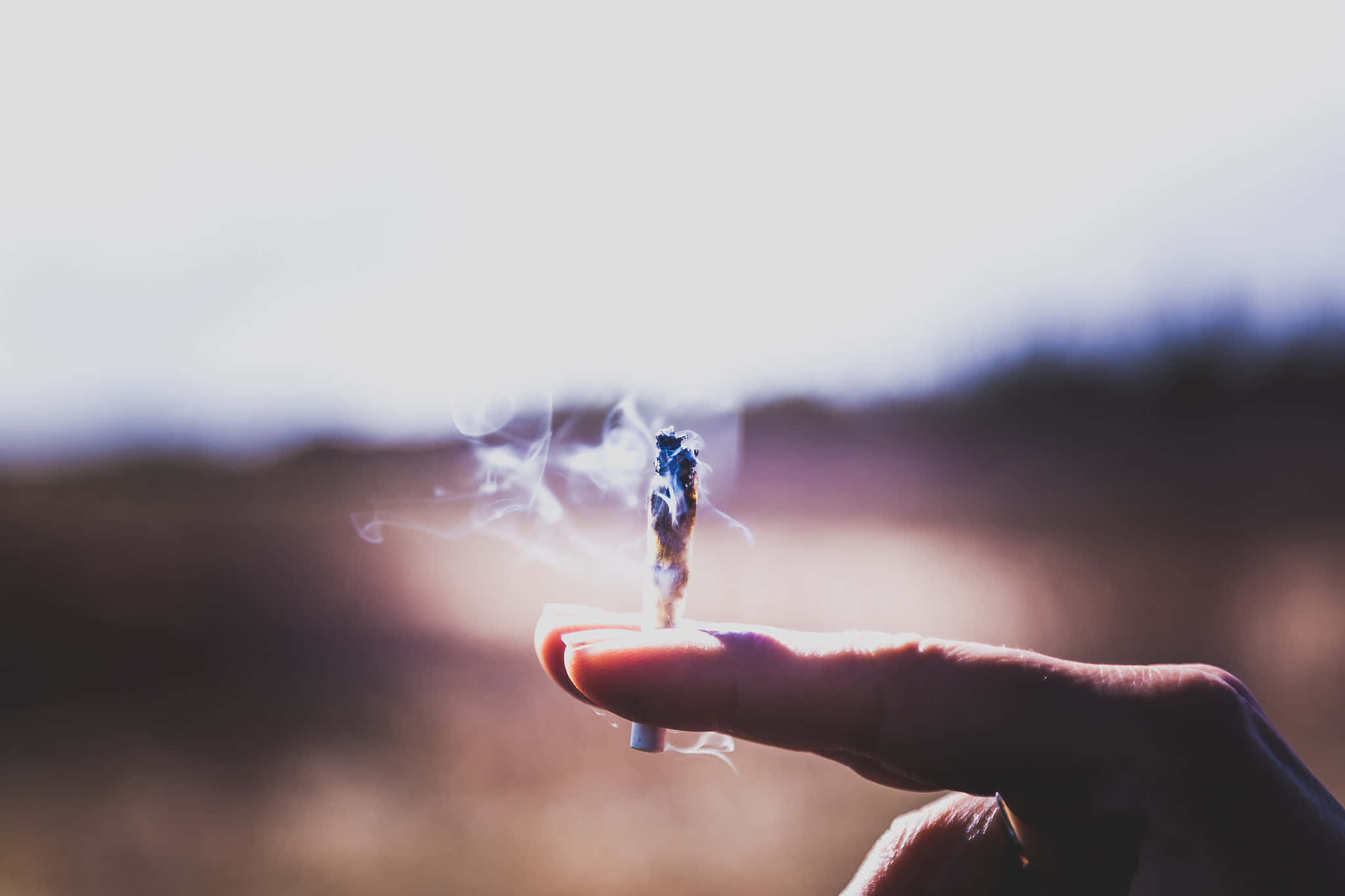 直立持煙卷 壁紙 — 一隻手拿著一支點燃的大麻煙，煙霧在模糊的戶外背景下裊裊升起。