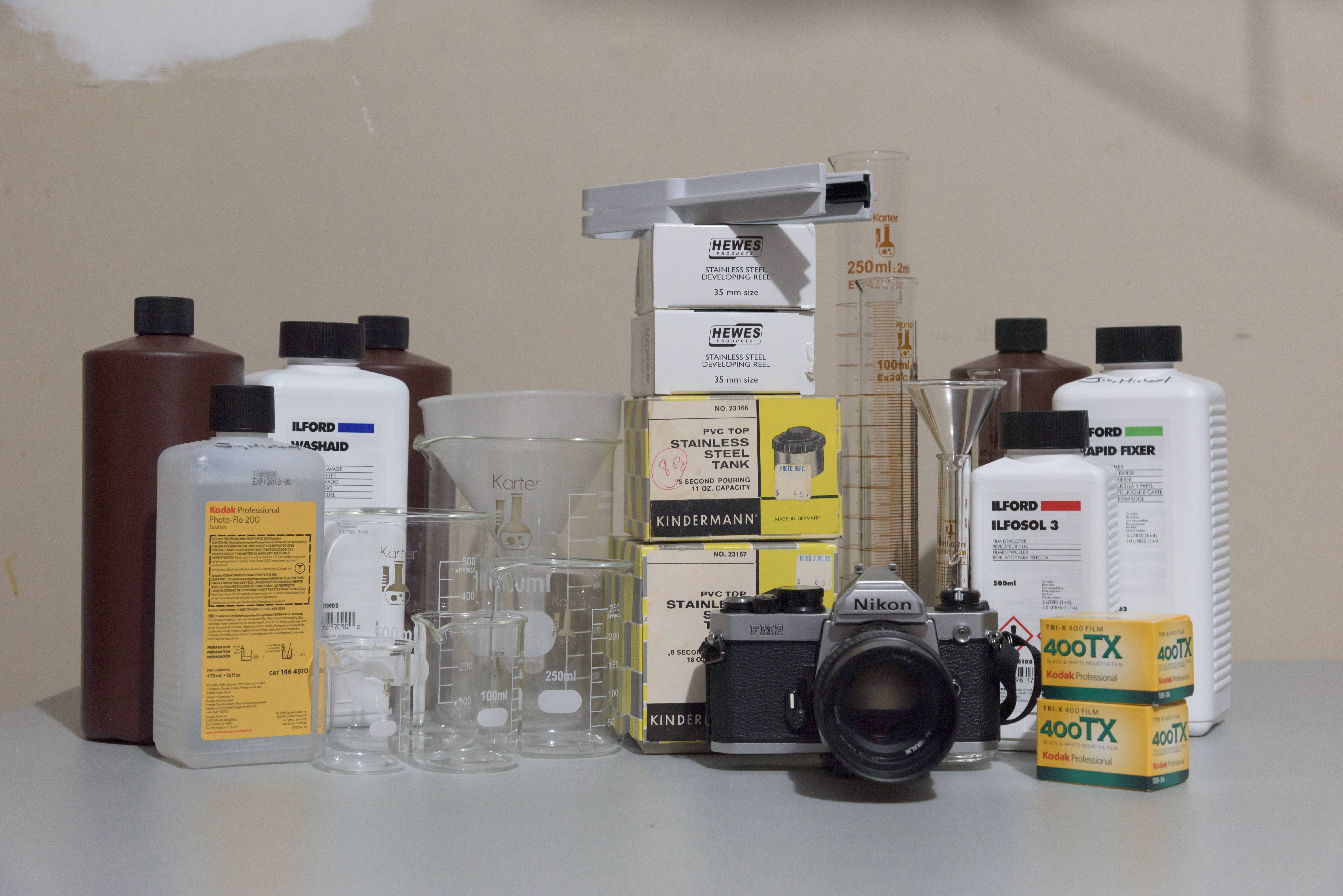 相機、燒杯與化學品 壁紙 — 一個復古菲林攝影設置，包括Nikon相機、化學品和玻璃器皿，排列在中性表面上。