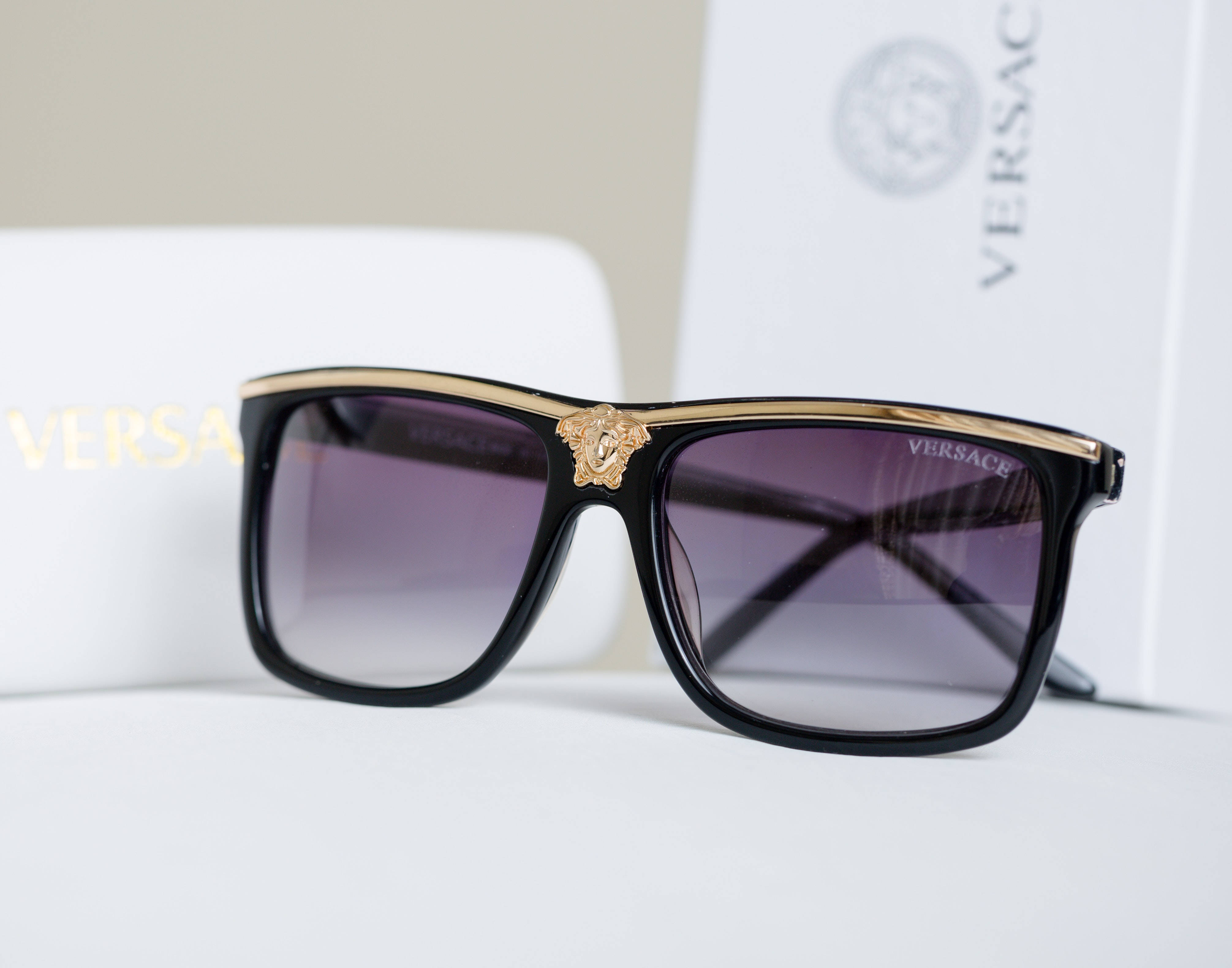 真品 Versace 太陽眼鏡 壁紙 — 帶有金色美杜莎標誌的黑色Versace太陽眼鏡大膽地放在白色表面上，旁邊是品牌包裝。