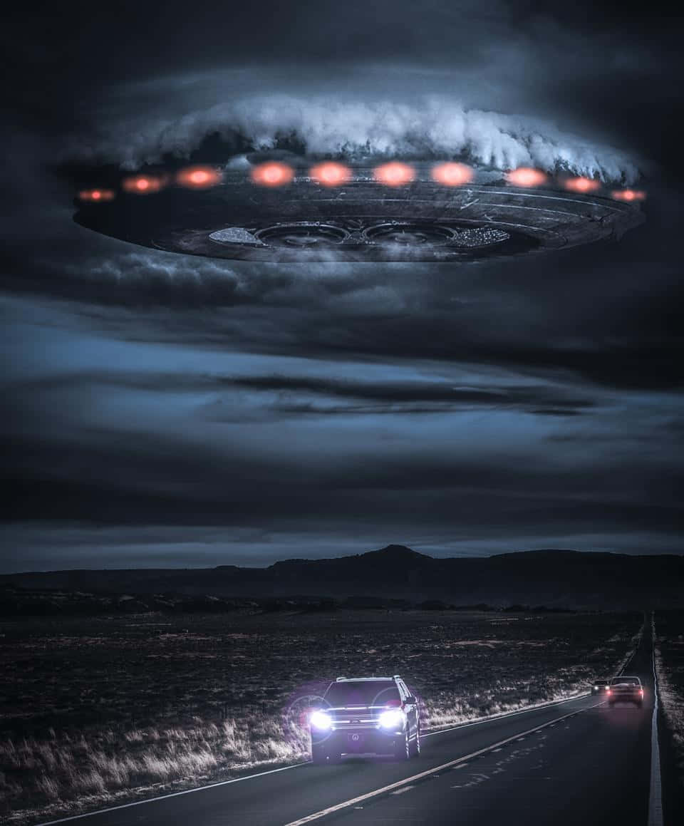 真實UFO飛船懸浮於路中汽車圖像 壁紙 — 一艘發光的UFO盤旋在黑暗的沙漠公路上方，汽車在暴風雨的天空下行駛在下方。
