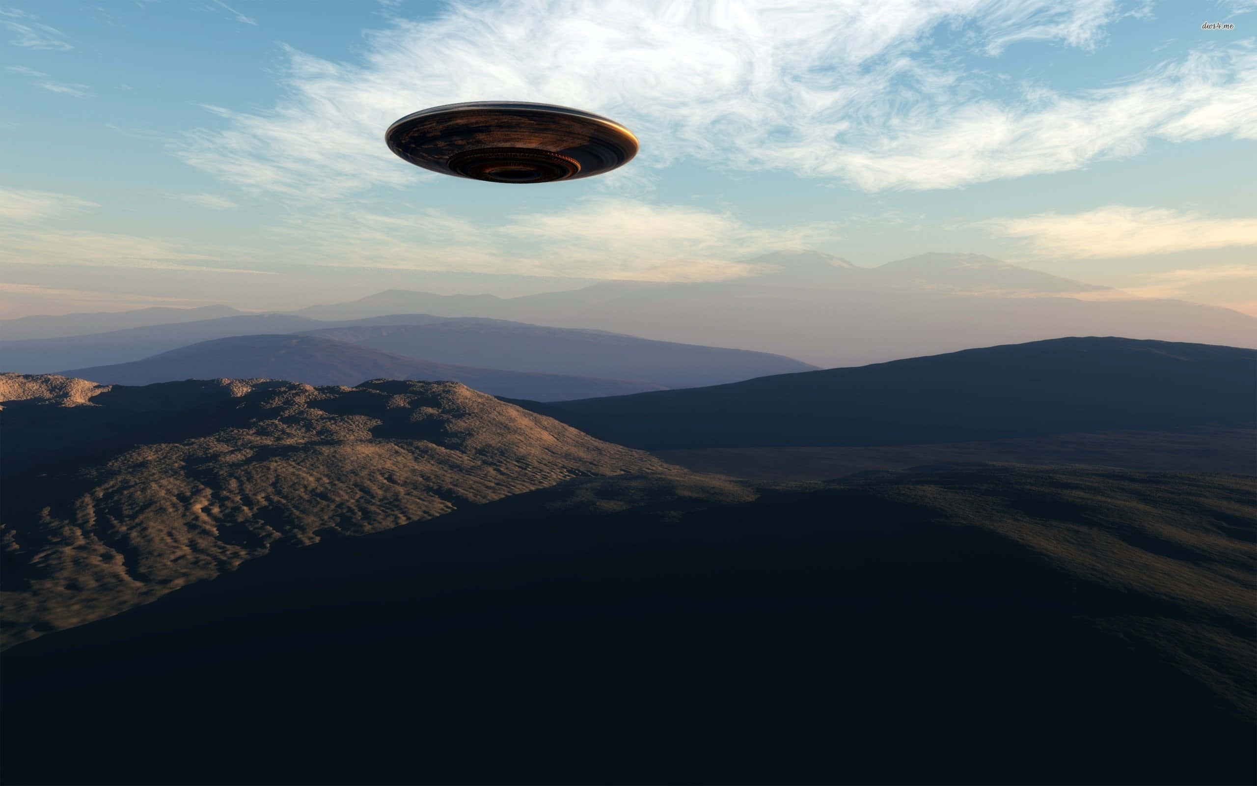 真實UFO飛越山巒天空圖像 壁紙 — 一個金屬UFO喺柔軟嘅日落天空下，帶住薄雲，盤旋喺崎嶇嘅山脈之上。