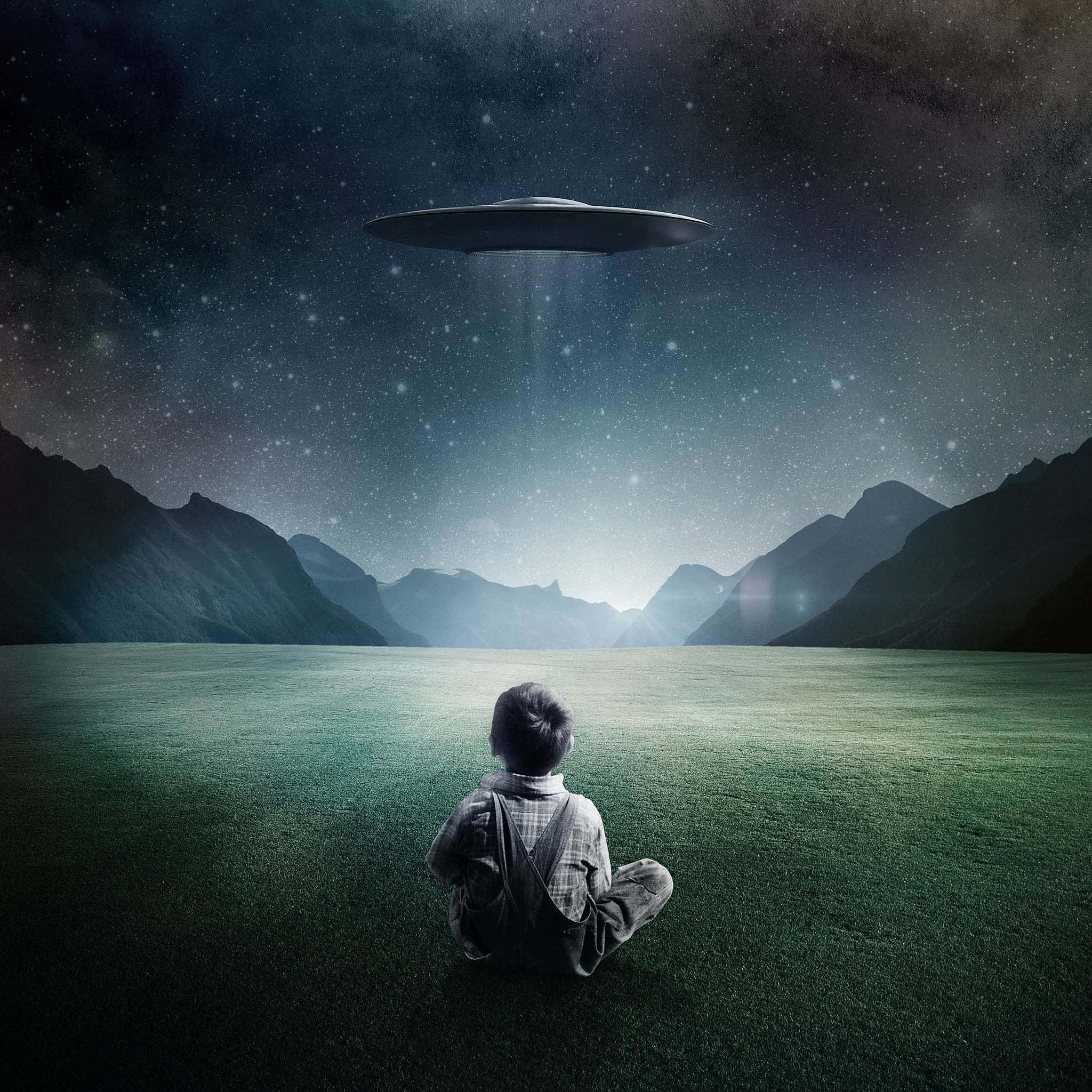 真實目擊：不明飛行物懸浮於天空 壁紙 — 一個孩子獨自坐在草地上，仰望在星空下遠山上方盤旋的發光UFO。