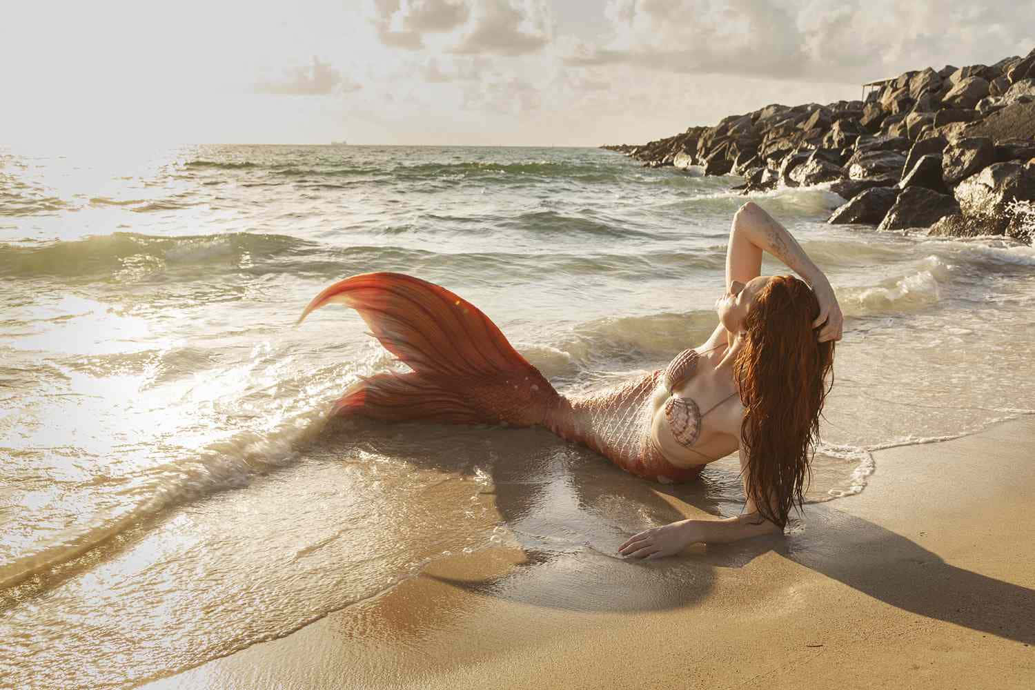 一條擁有橙色尾巴的美人魚在日落時分的海灘上休息。