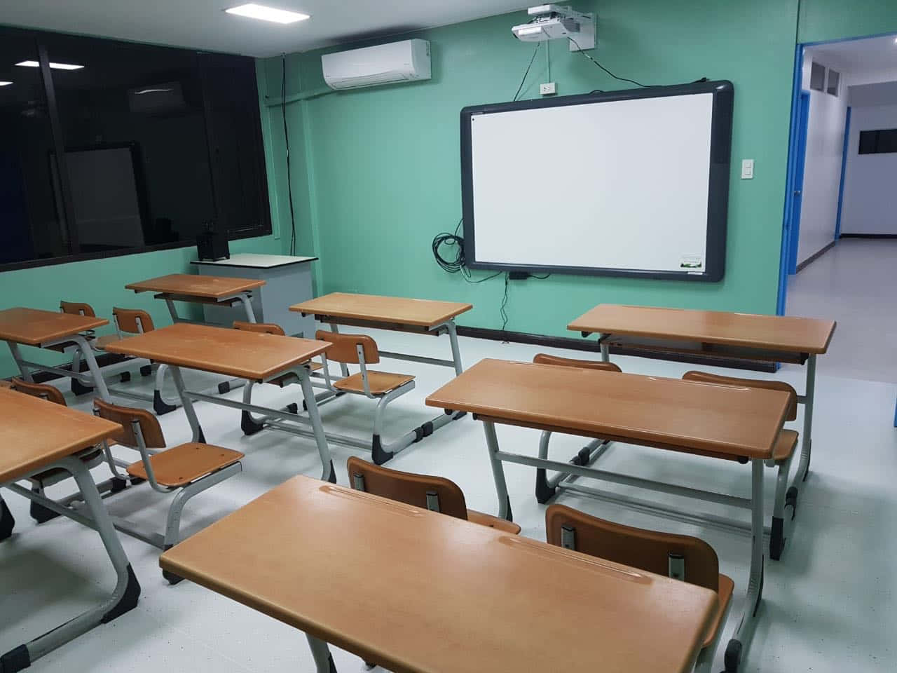 真實課堂中學習與成長 壁紙 — 空無一人的教室，有木製書桌、椅子，以及螢光燈下的大型白板。
