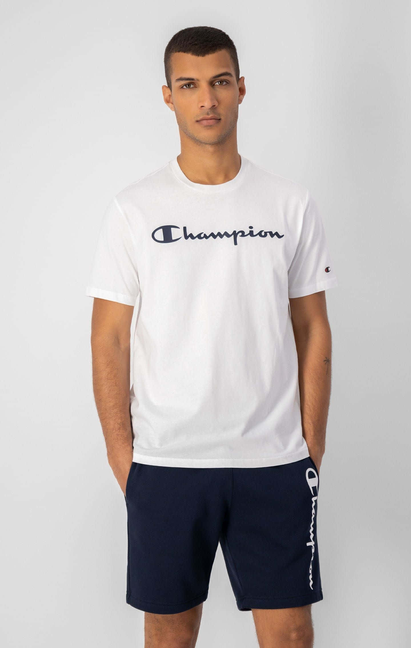 真正的冠軍風采 壁紙 — 一名身穿白色Champion T恤和深藍色短褲的男子自信地站在純灰色背景前。