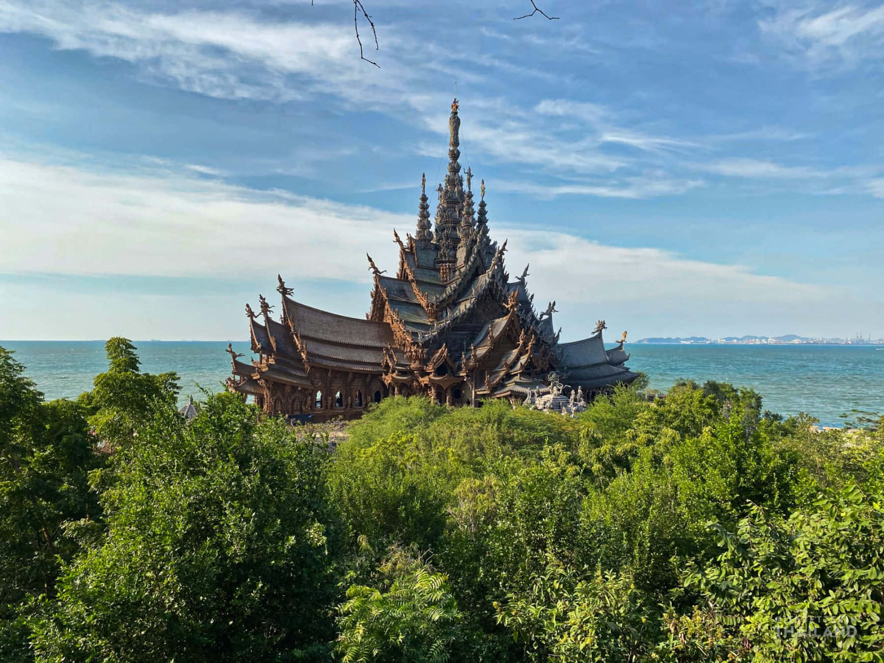 真理聖殿從森林中探出 壁紙 — 一座雕刻精美的木製寺廟從茂密的綠植中升起，俯瞰著廣闊的藍色大海，天空部分多雲。