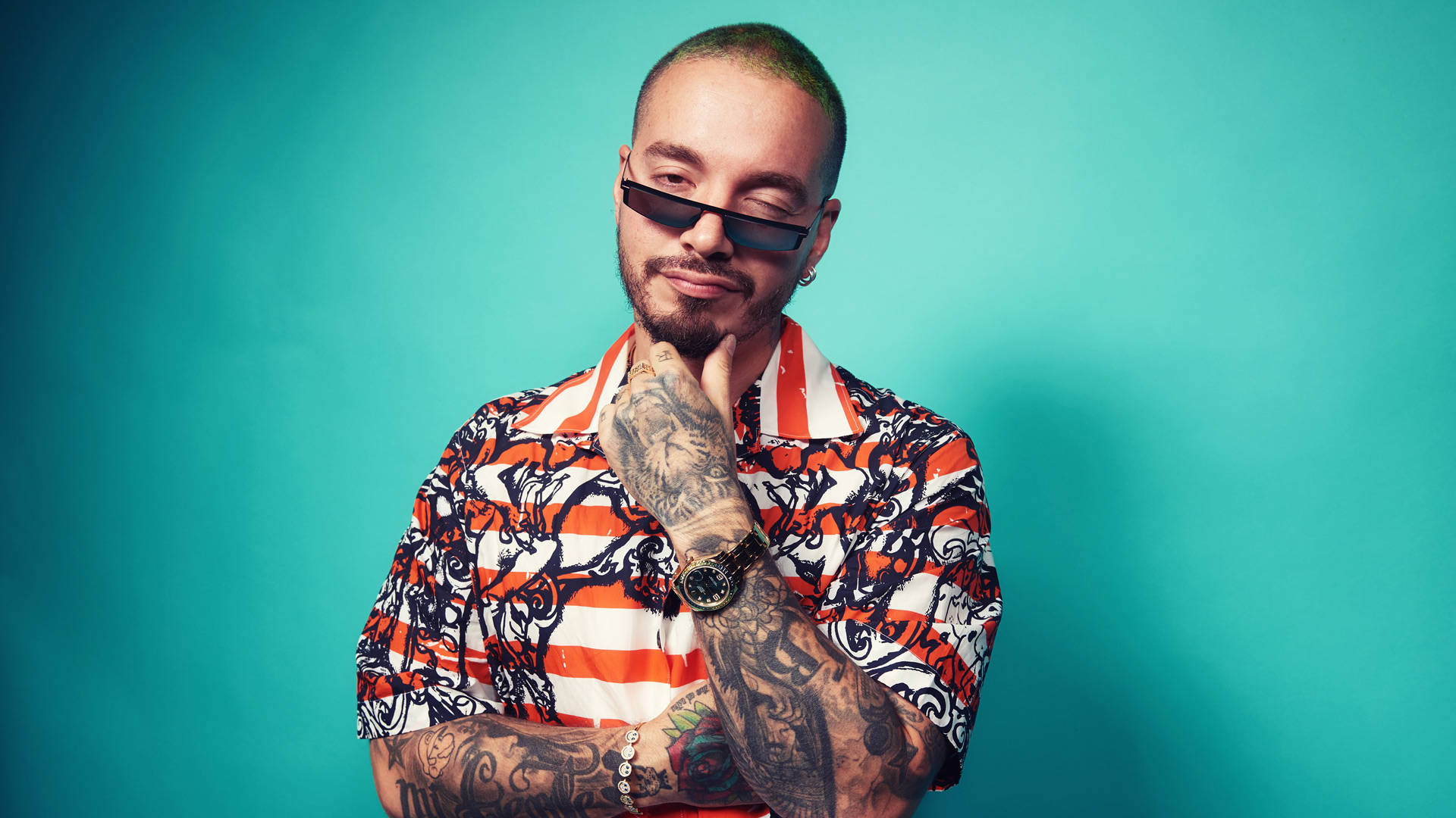 眨眼的 J Balvin 壁紙 — 一個時尚的紋身男子穿著大膽圖案襯衫，在純青色背景前自信地擺姿勢。