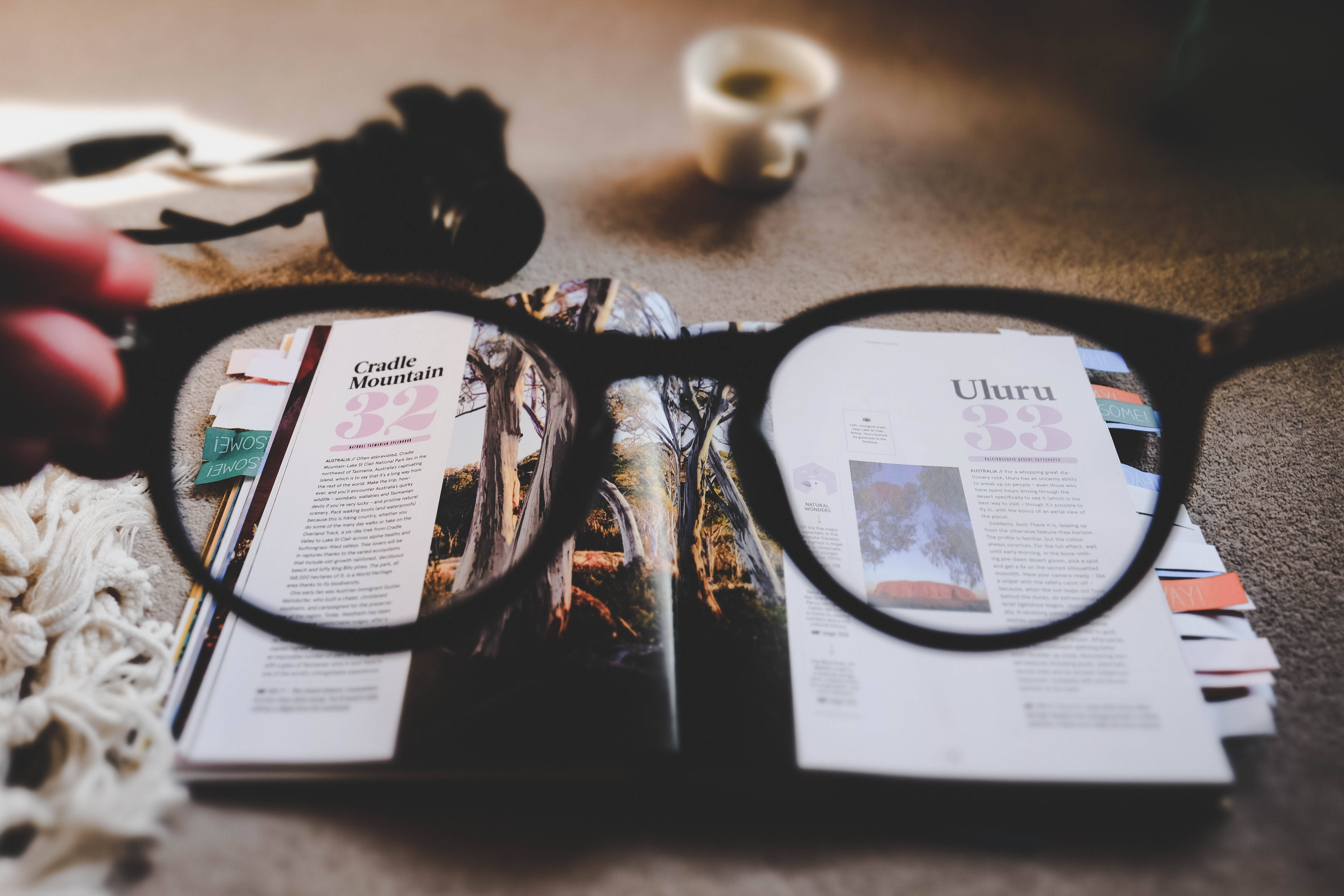 眼鏡放大鏡下的雜誌 壁紙 — 一隻手拿著黑色框眼鏡，放在打開的旅遊雜誌上，雜誌上有搖籃山和烏魯魯，附近有一杯咖啡。