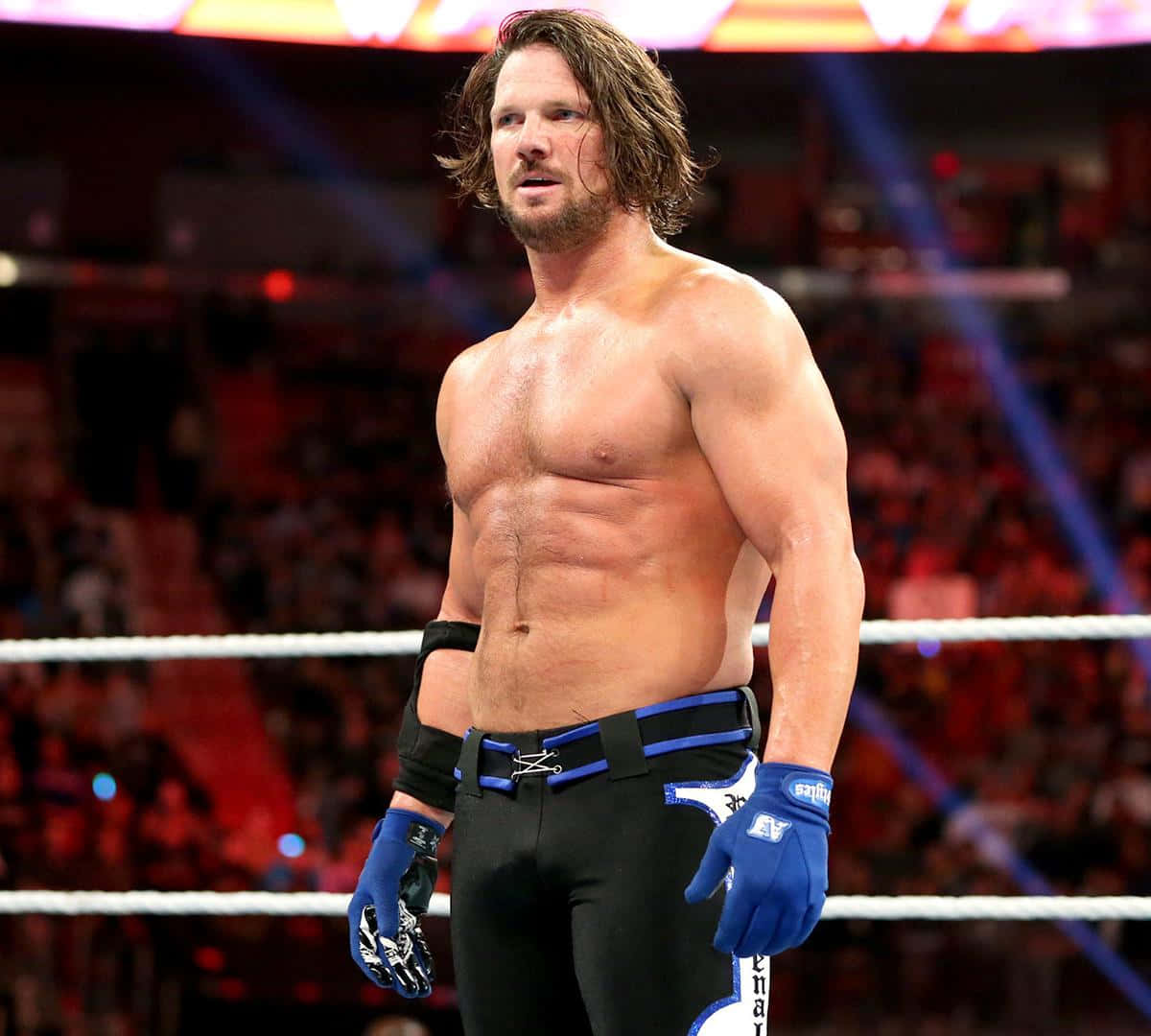 知名摔角手 AJ Styles 壁紙 — 一位肌肉發達的長髮摔角手站在擂台上，穿著黑色短褲和藍色手套，在明亮的場館燈光下。