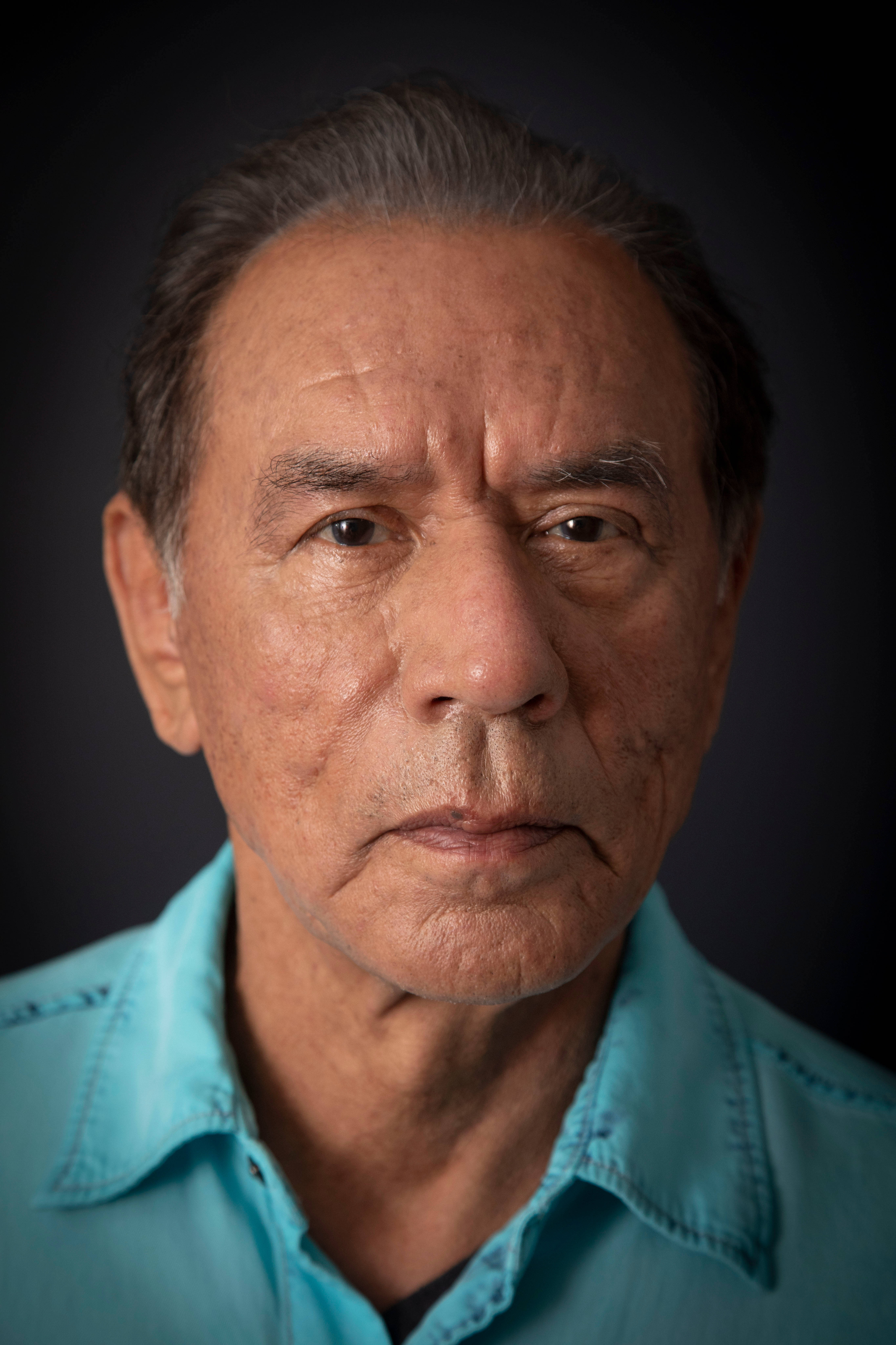 知名演員 Wes Studi 畫像 壁紙 — 一位灰白頭髮、滿臉皺紋的長者身穿青綠色襯衫，在深色背景下的特寫肖像。