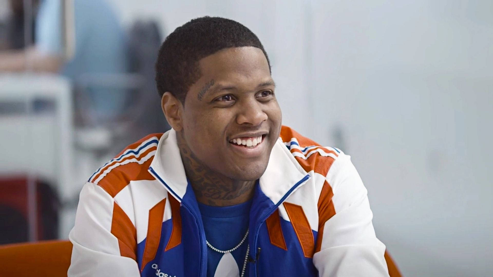 知名說唱歌手 Lil Durk 壁紙 — 一位微笑的男子穿著白色、橙色和藍色的Adidas運動外套，露出頸部紋身，坐在室內。
