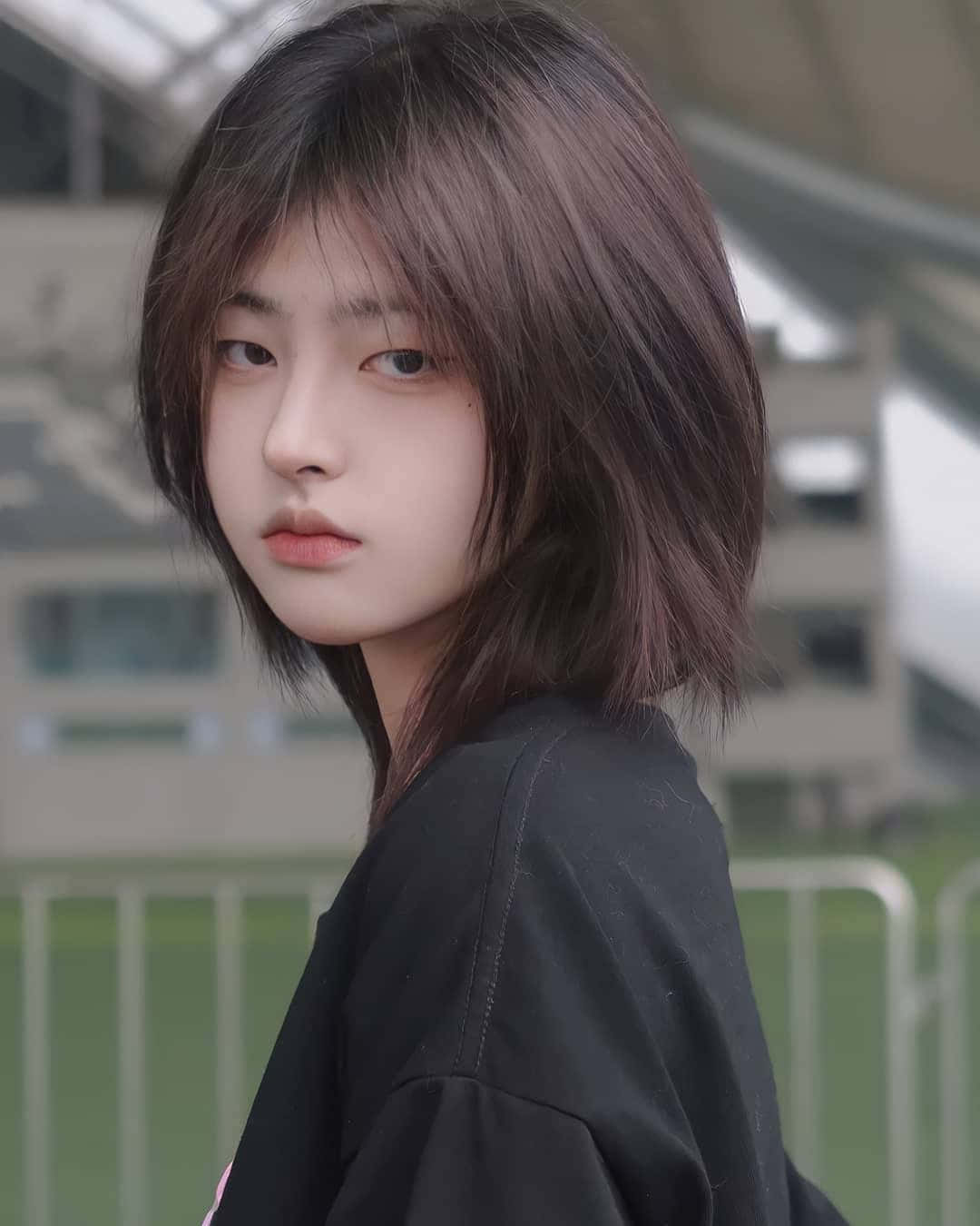 短髮造型女孩照片 壁紙 — 一位短黑髮的年輕女性，穿著黑色上衣，在模糊的體育場背景下微微轉頭。