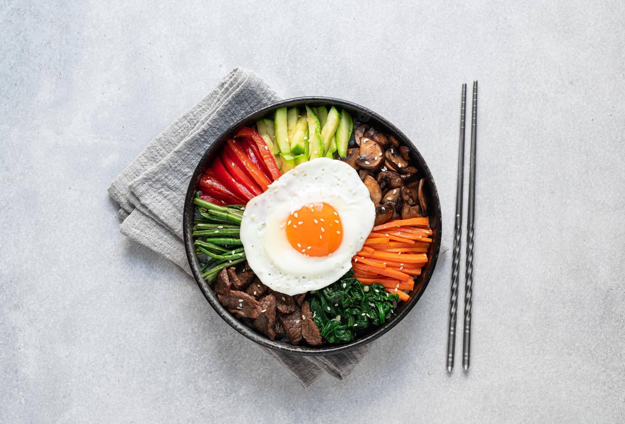 石鍋拌飯擺放在石檯上 壁紙 — 一碗色彩繽紛的韓國石鍋拌飯，上面放著太陽蛋，放在灰色餐巾上，旁邊有筷子。