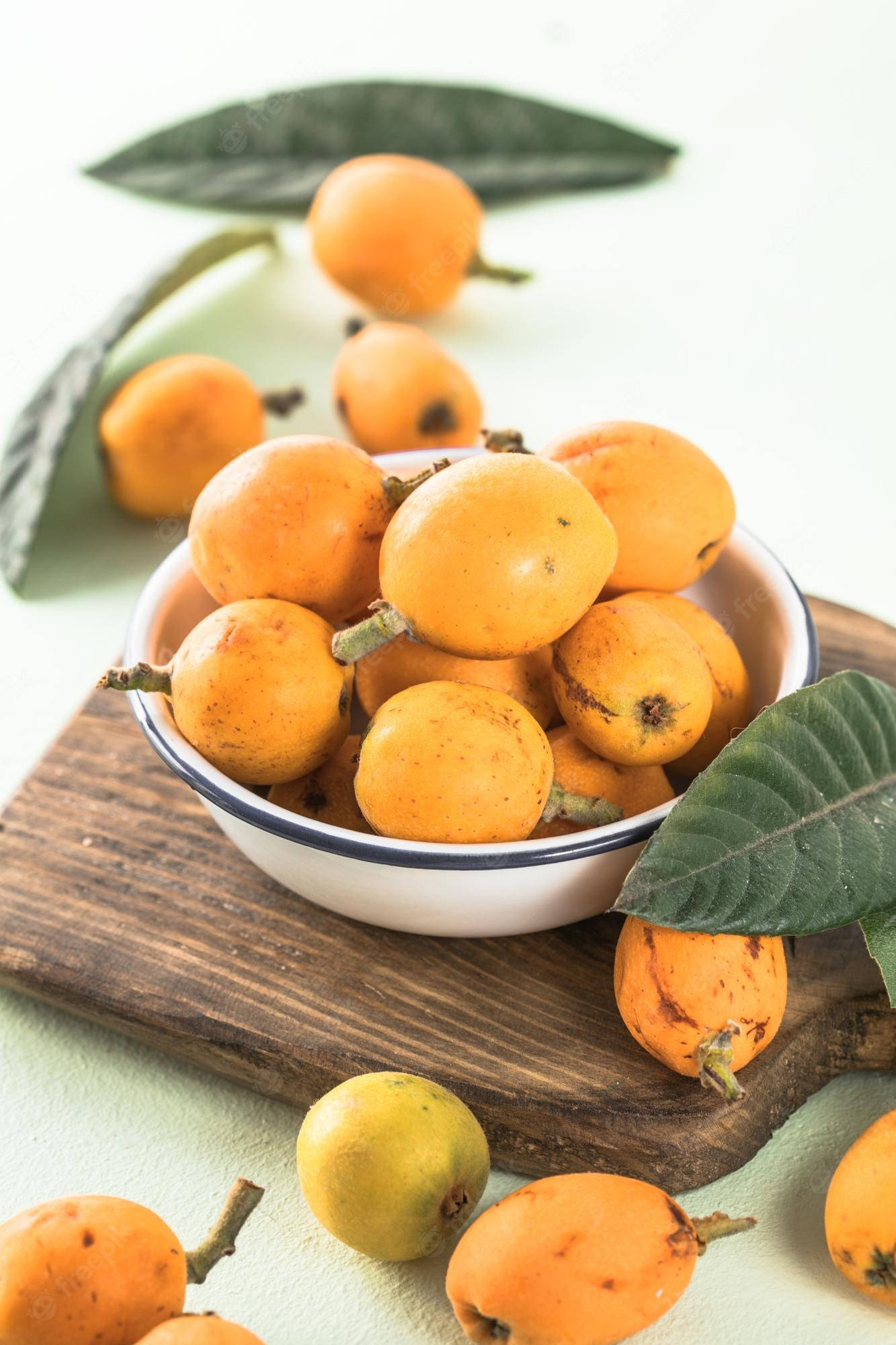 砧板上的新鮮枇杷果 壁紙 — 一個白色碗裝滿成熟金黃色枇杷，放在一塊鄉村風格木板上，周圍有新鮮綠葉和散落的水果。