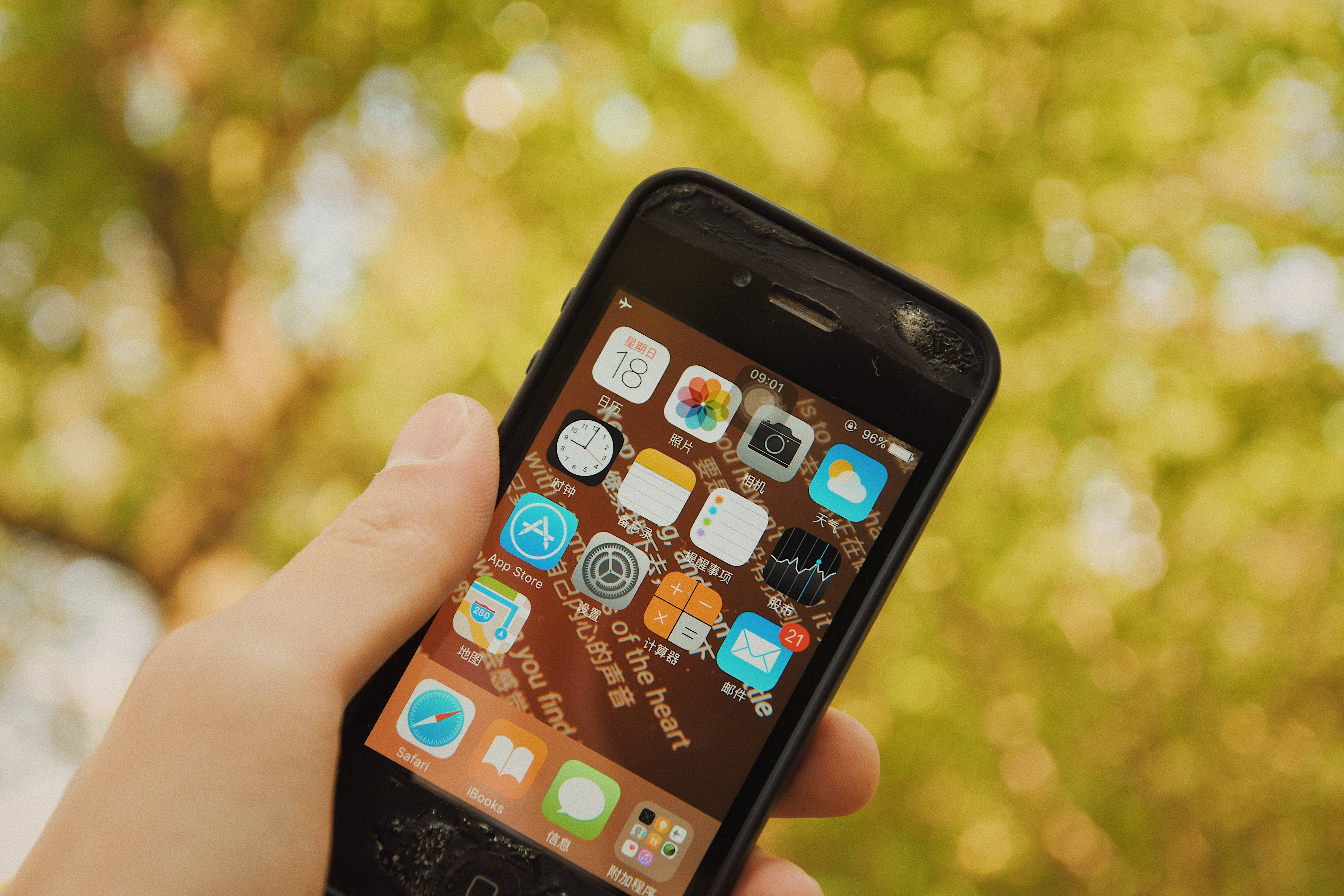 破碎的iOS iPhone 壁紙 — 一隻手拿著一部破裂的智能手機，顯示色彩繽紛的應用程式網格，背景是柔和模糊的綠色樹葉。