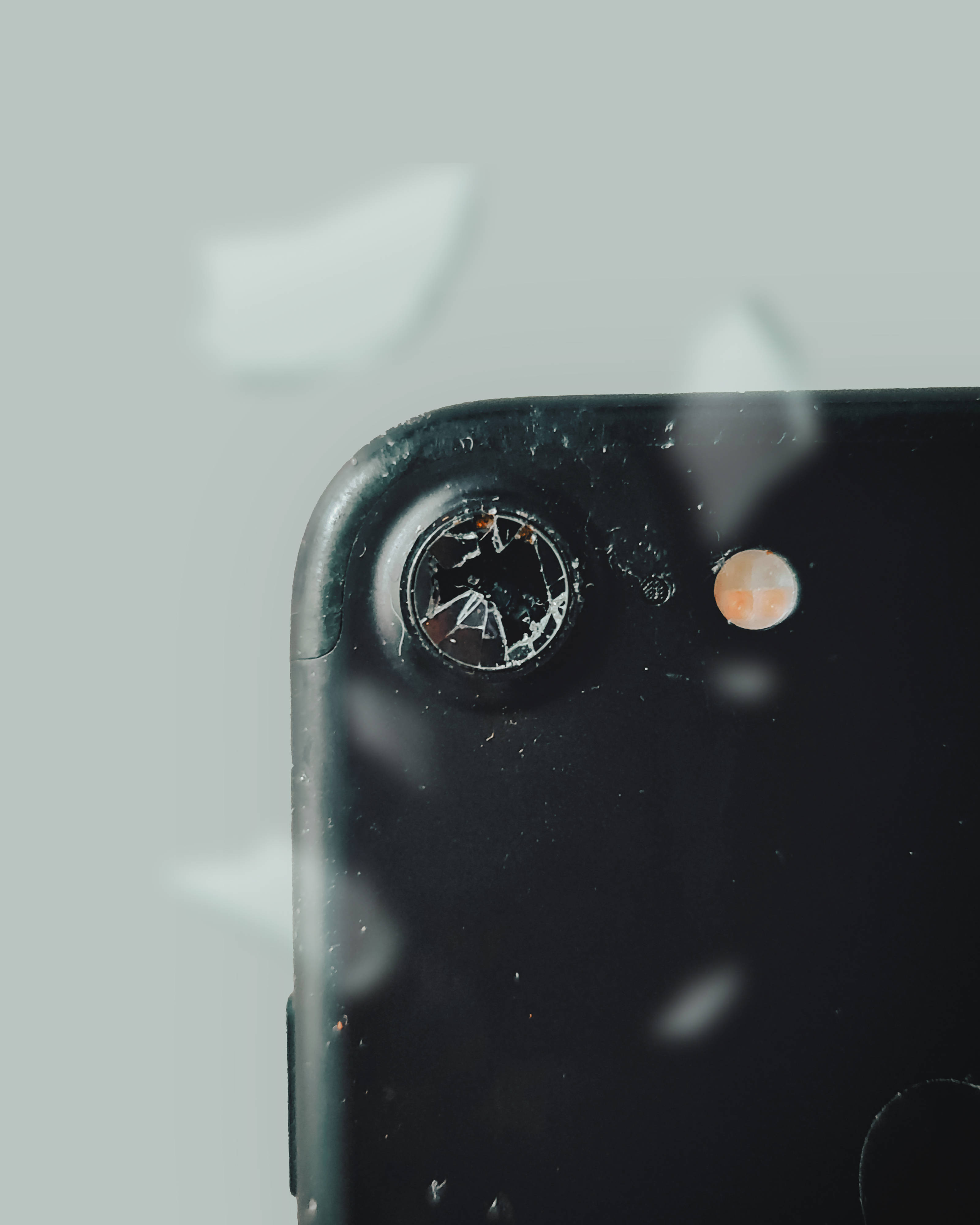 碎裂手機鏡頭 壁紙 — 淡灰色背景上，黑色智能手機的特寫，鏡頭破裂，玻璃碎片漂浮。
