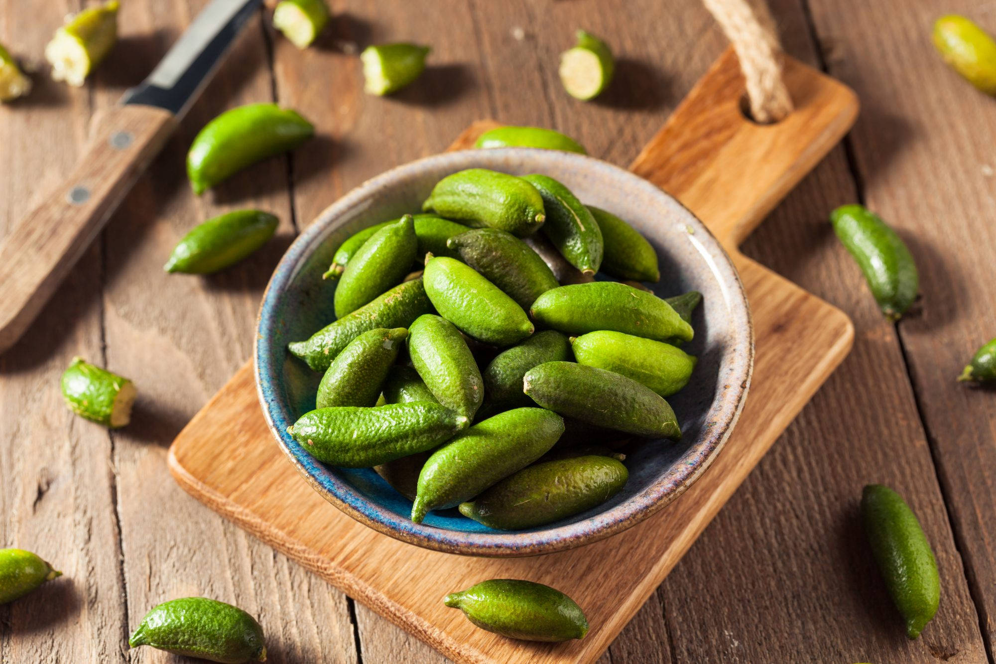 碗中盛放的指 lime 果 壁紙 — 一個質樸的陶瓷碗裝滿了鮮綠色的金橘，放在木製砧板上，周圍散落著切半的金橘和一把刀，擱在經風化的木桌上。