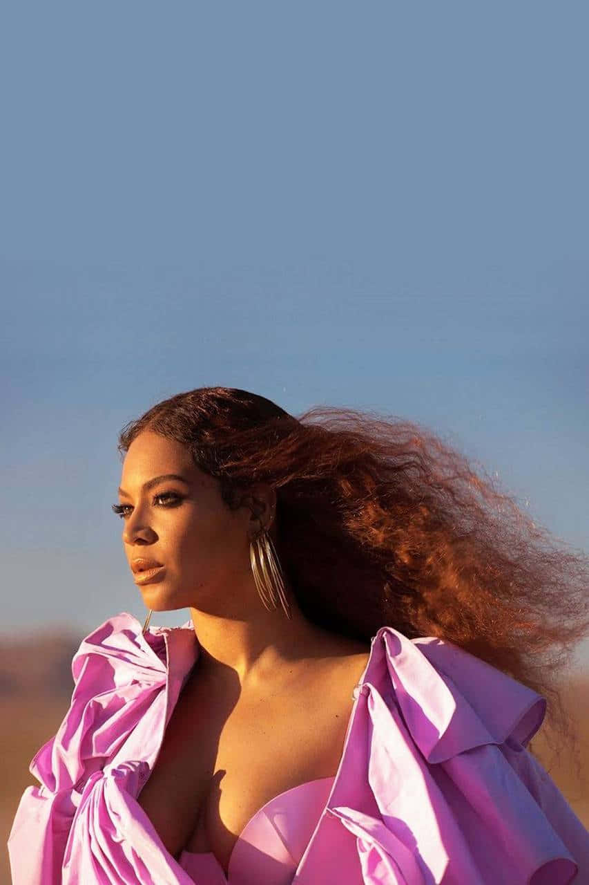 碧昂絲 Knowles 舞台光芒萬丈 壁紙 — Beyoncé 站在沙漠景觀中，穿著充滿活力的粉紅色荷葉邊連衣裙，風吹過她長長的捲髮，在清澈的藍天下。