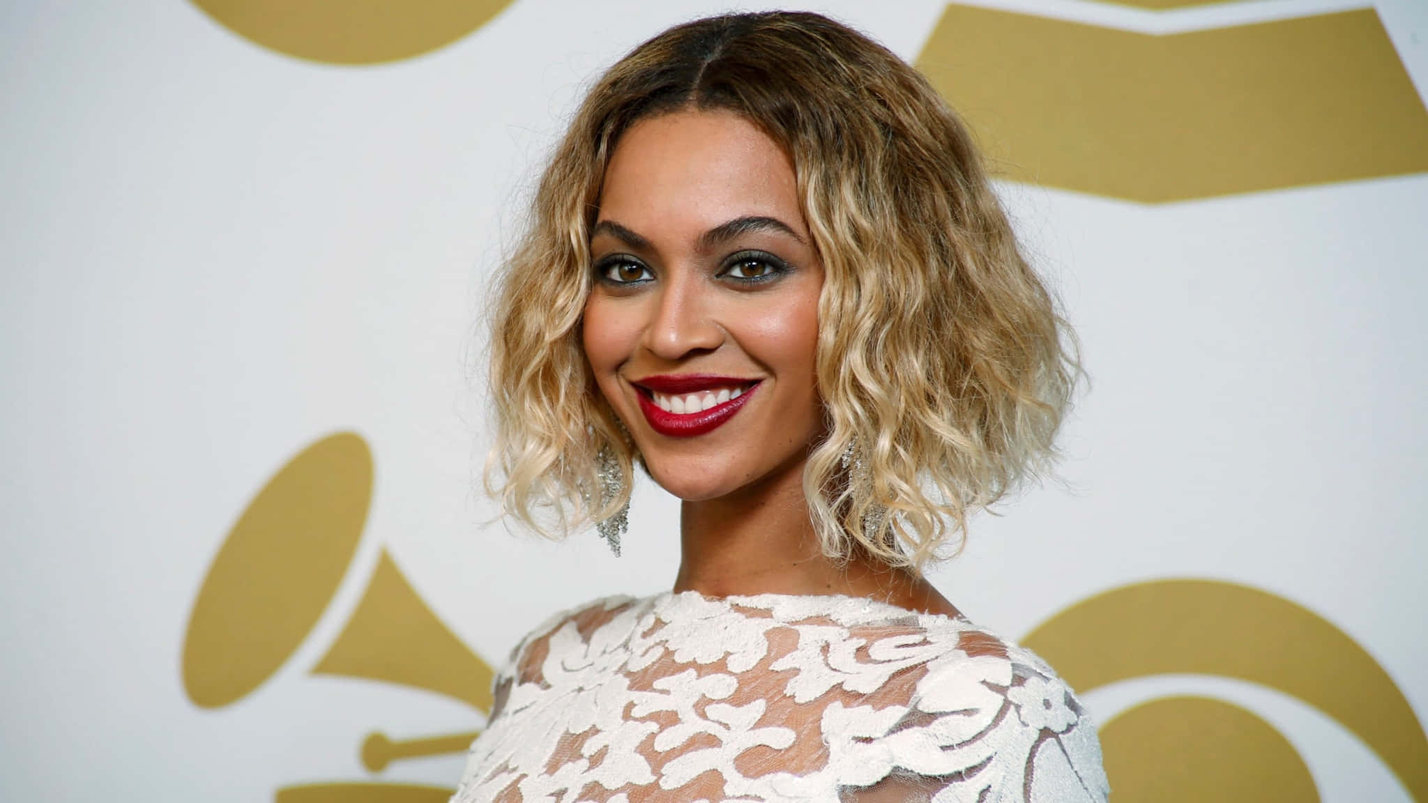 碧昂絲——流行音樂傳奇 壁紙 — Beyoncé 在金色葛萊美標誌背景下穿著白色蕾絲連衣裙燦爛微笑。