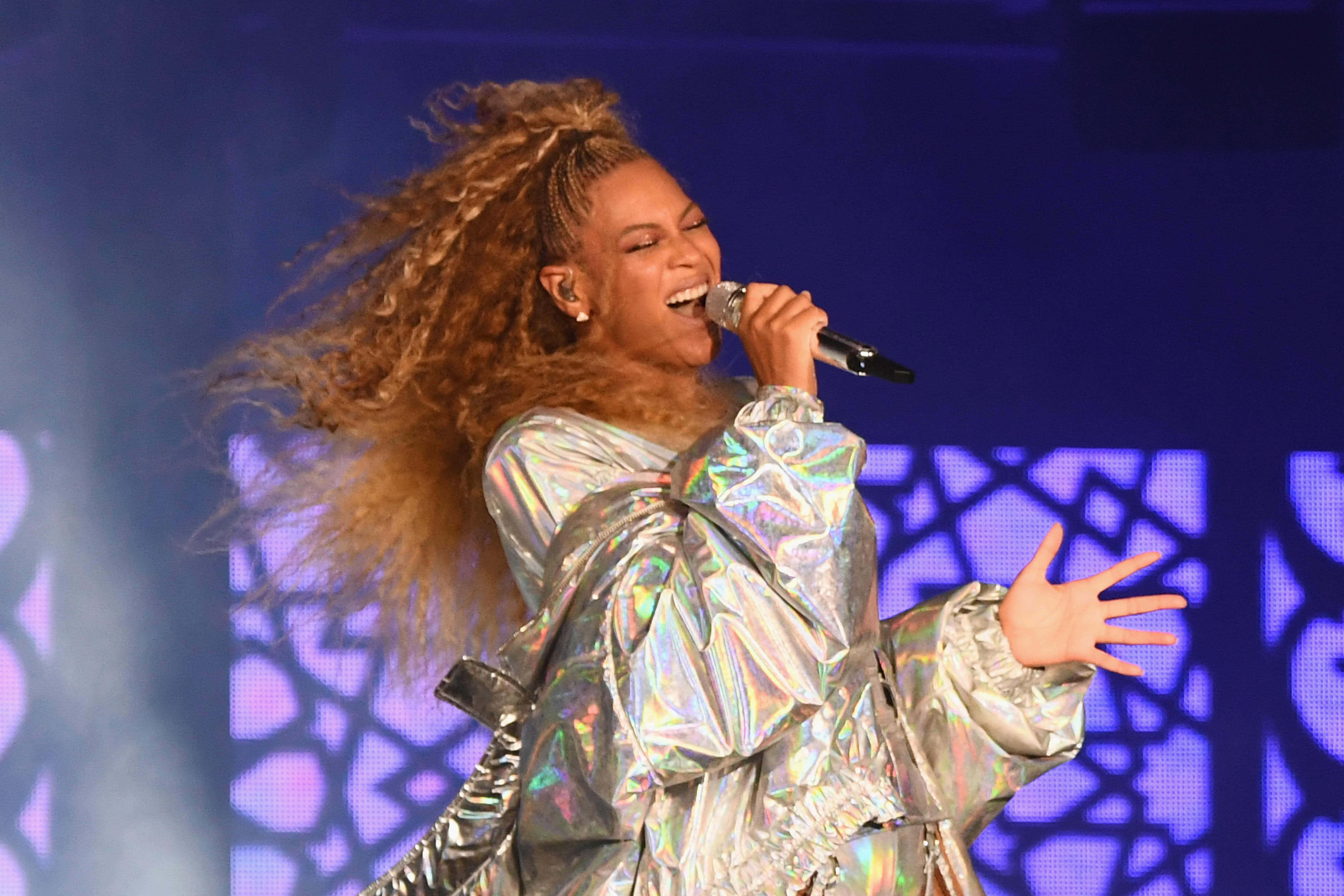 碧昂絲於2011年在羅斯蘭舞廳演唱熱門單曲《Love on Top》 壁紙 — Beyoncé 在紫色圖案背景前，身穿閃爍銀色服裝，長髮飄逸，在舞台上熱情表演。